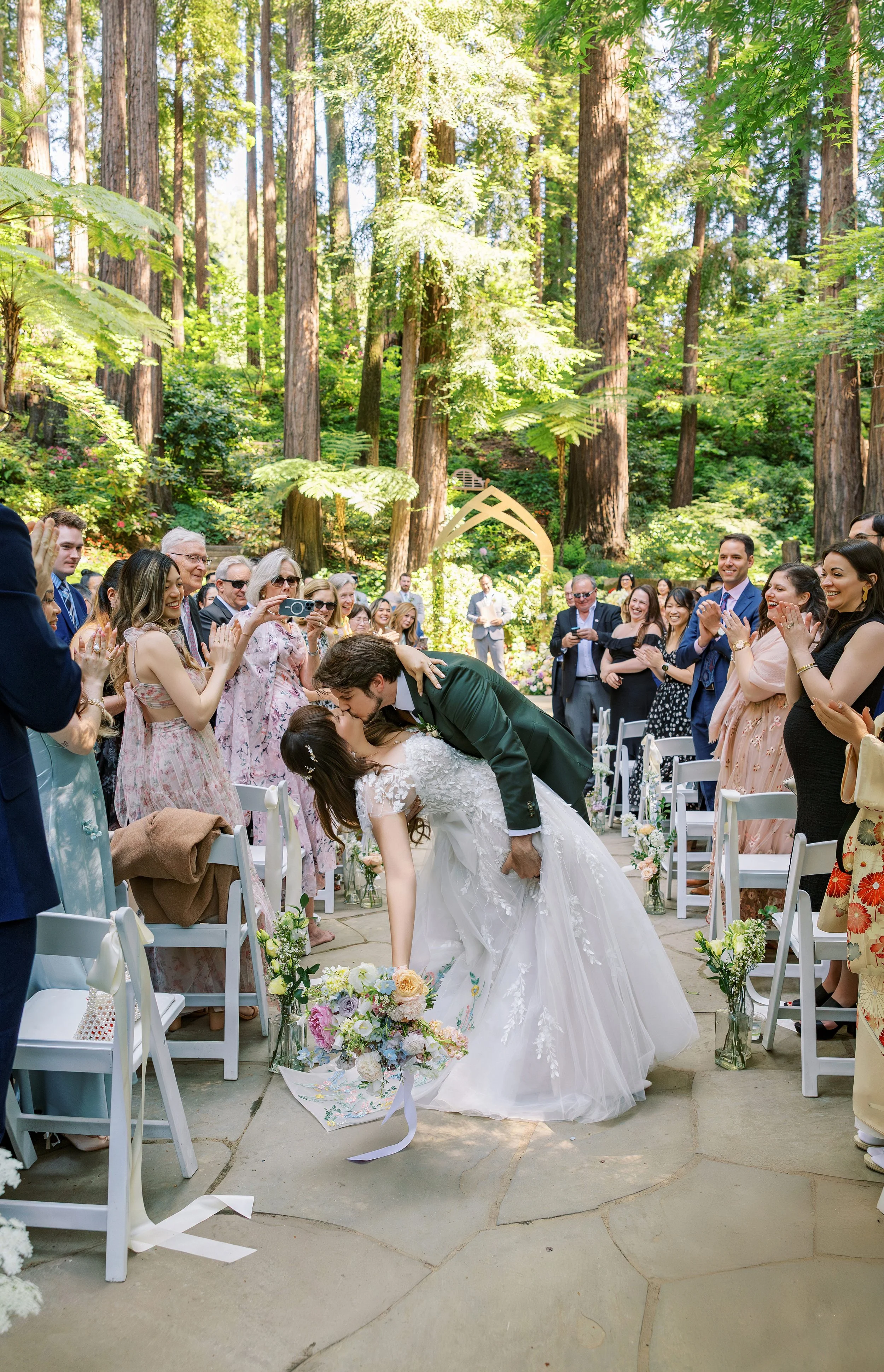 A bride and groom share a kiss at their outdoor wedding ceremony surrounded by friends and family in a forest setting.