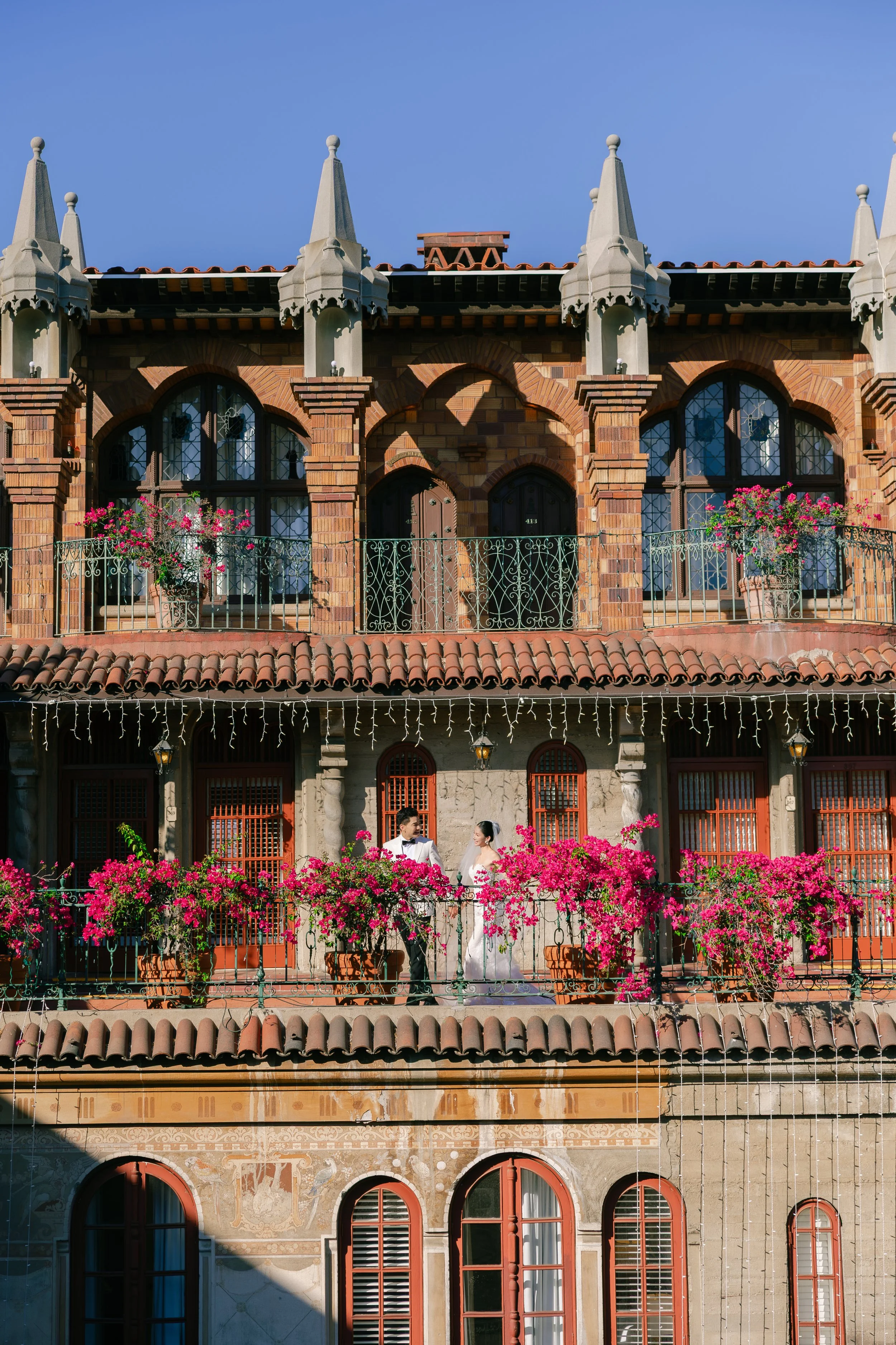A bride and groom standing on a balcony decorated with pink flowers, in front of an ornate building with arched windows and balconies, under a clear blue sky.