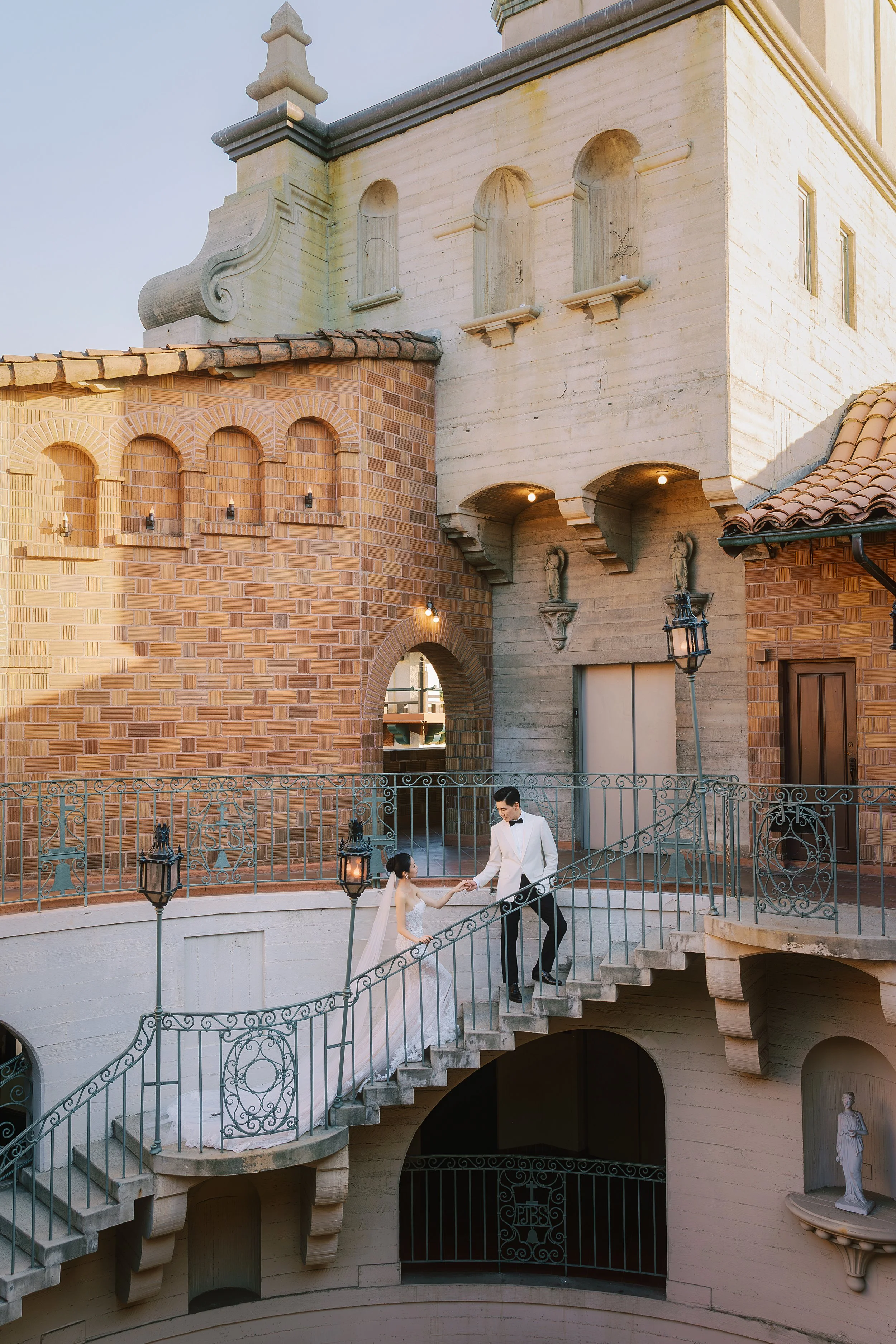 A bride and groom in wedding attire holding hands on an outdoor staircase of an ornate building with architectural details, statues, and decorative lamps.