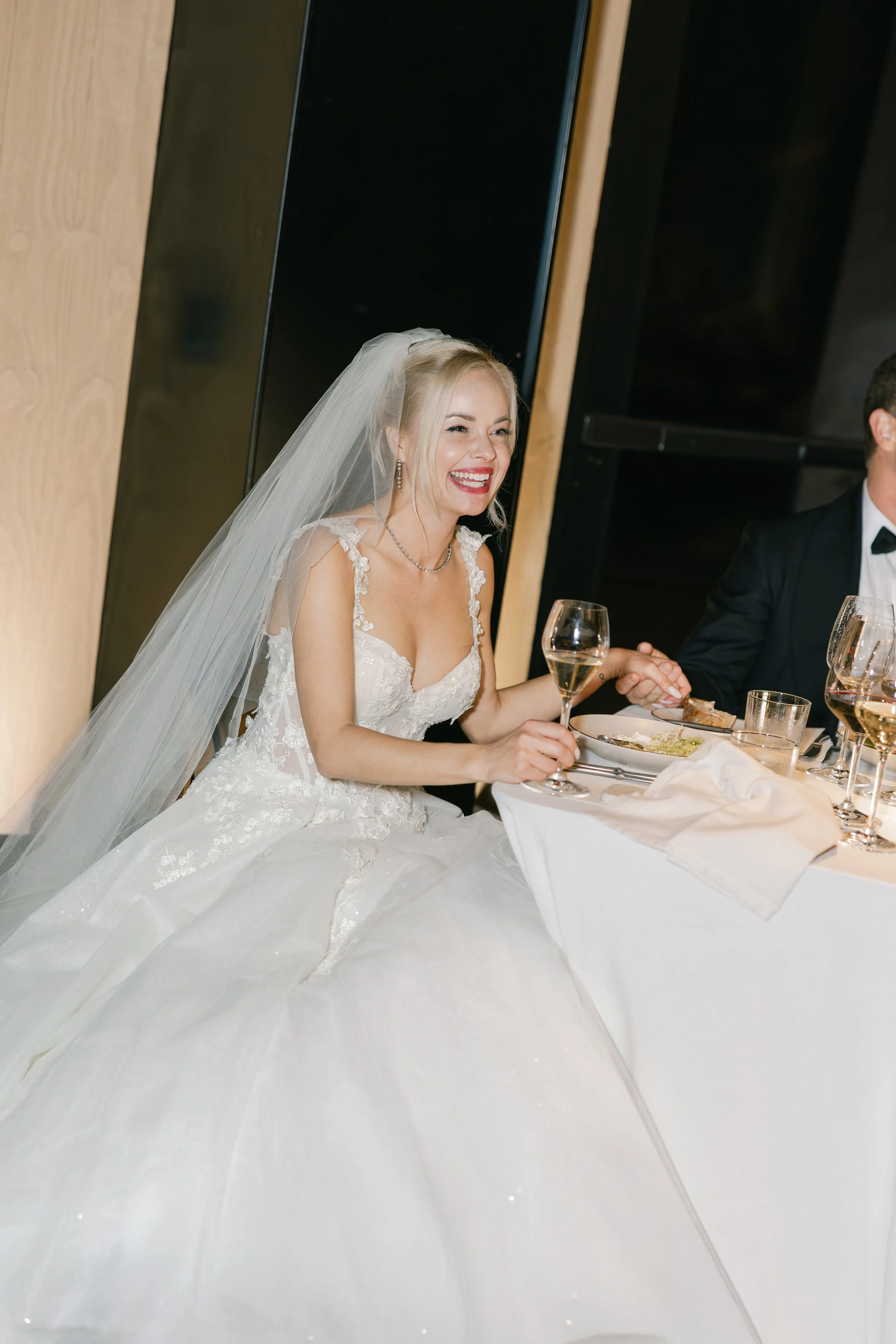 A bride in a white wedding gown laughing at a wedding reception, sitting at a table with a glass of champagne.