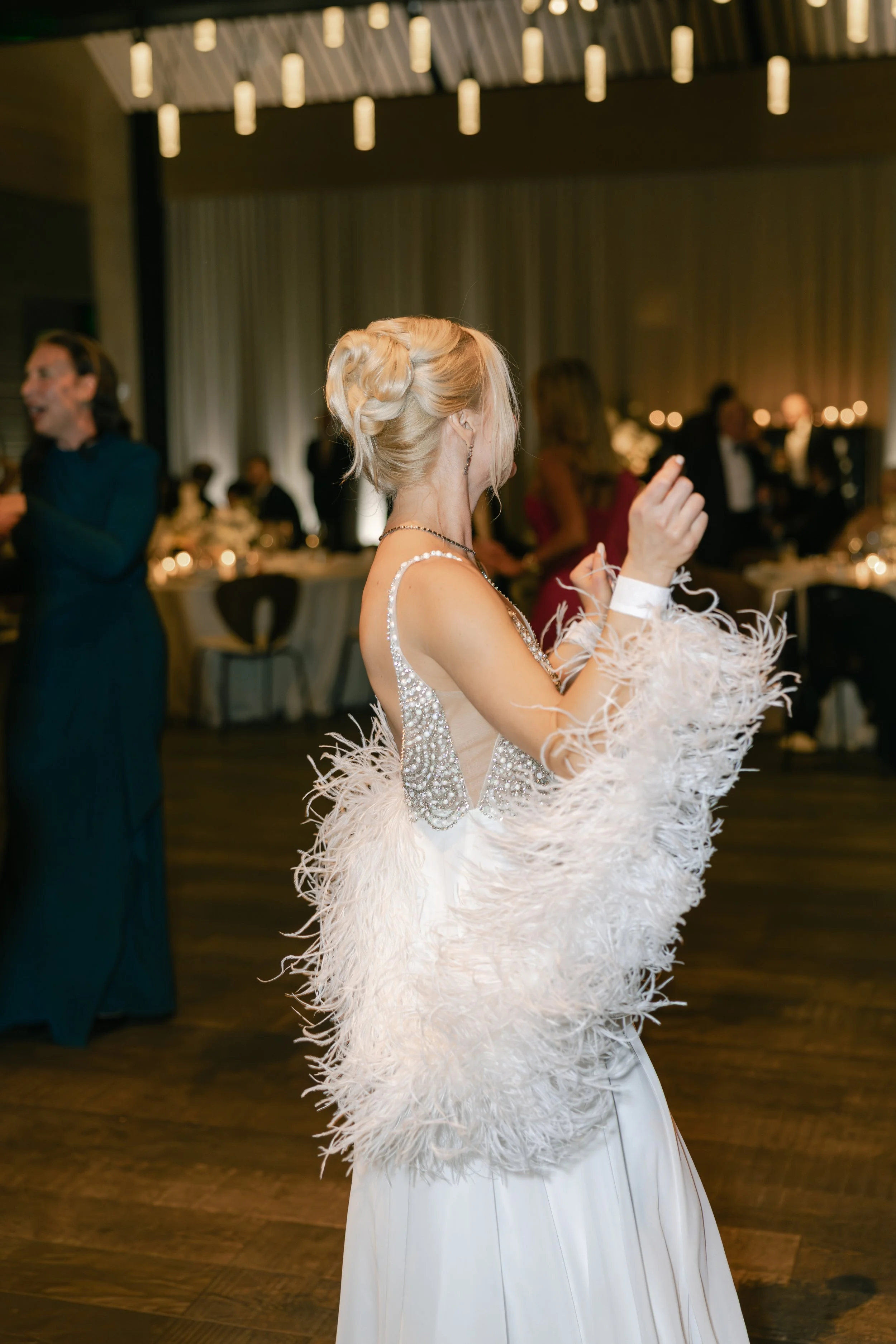 Woman in an elegant, beaded dress with feathered shawl dancing at a formal event, with other formally dressed guests and table settings in the background.