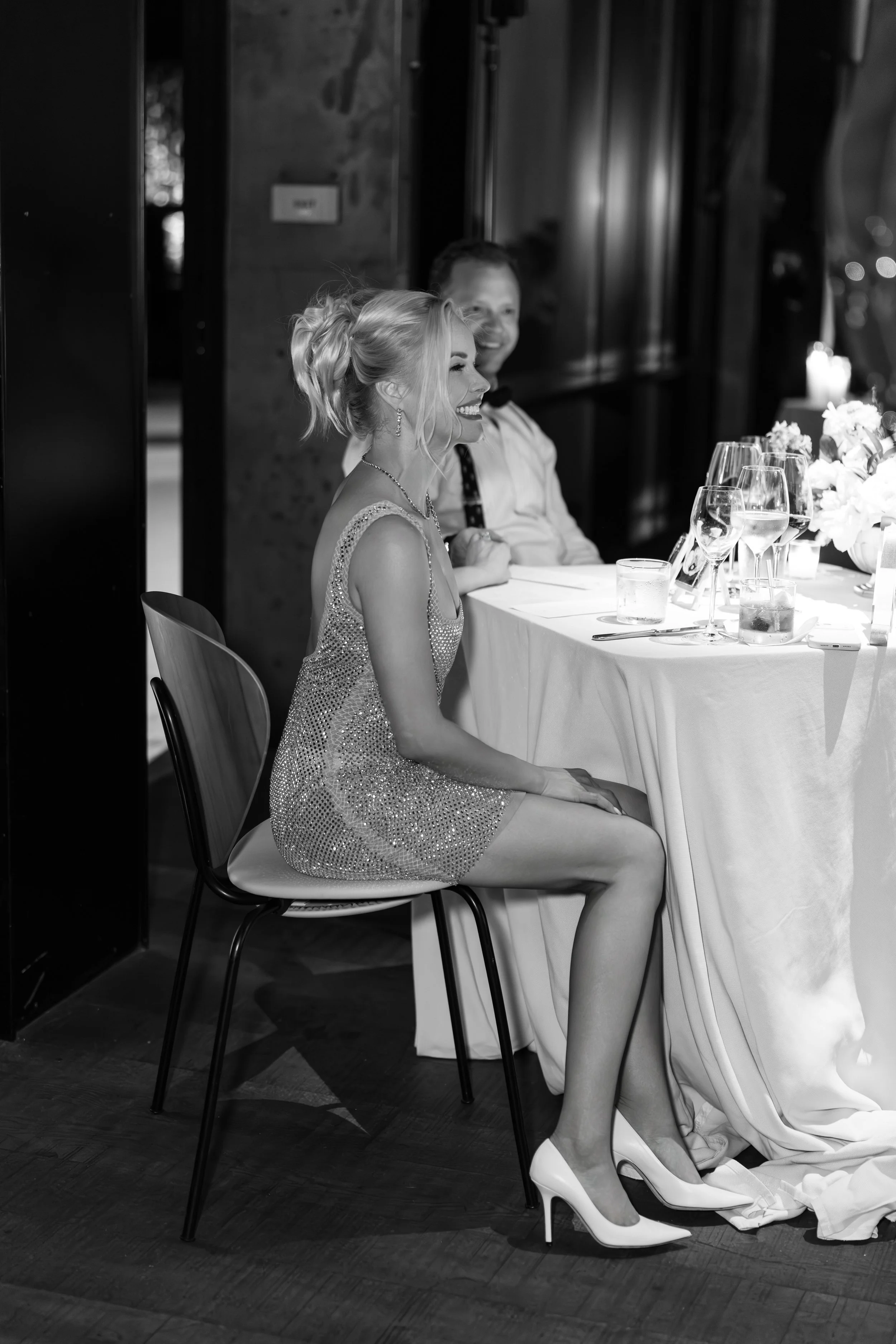 A woman in a sparkly dress and high heels sitting at a banquet table, smiling, with a man in a white shirt beside her, both appearing happy at a formal event.