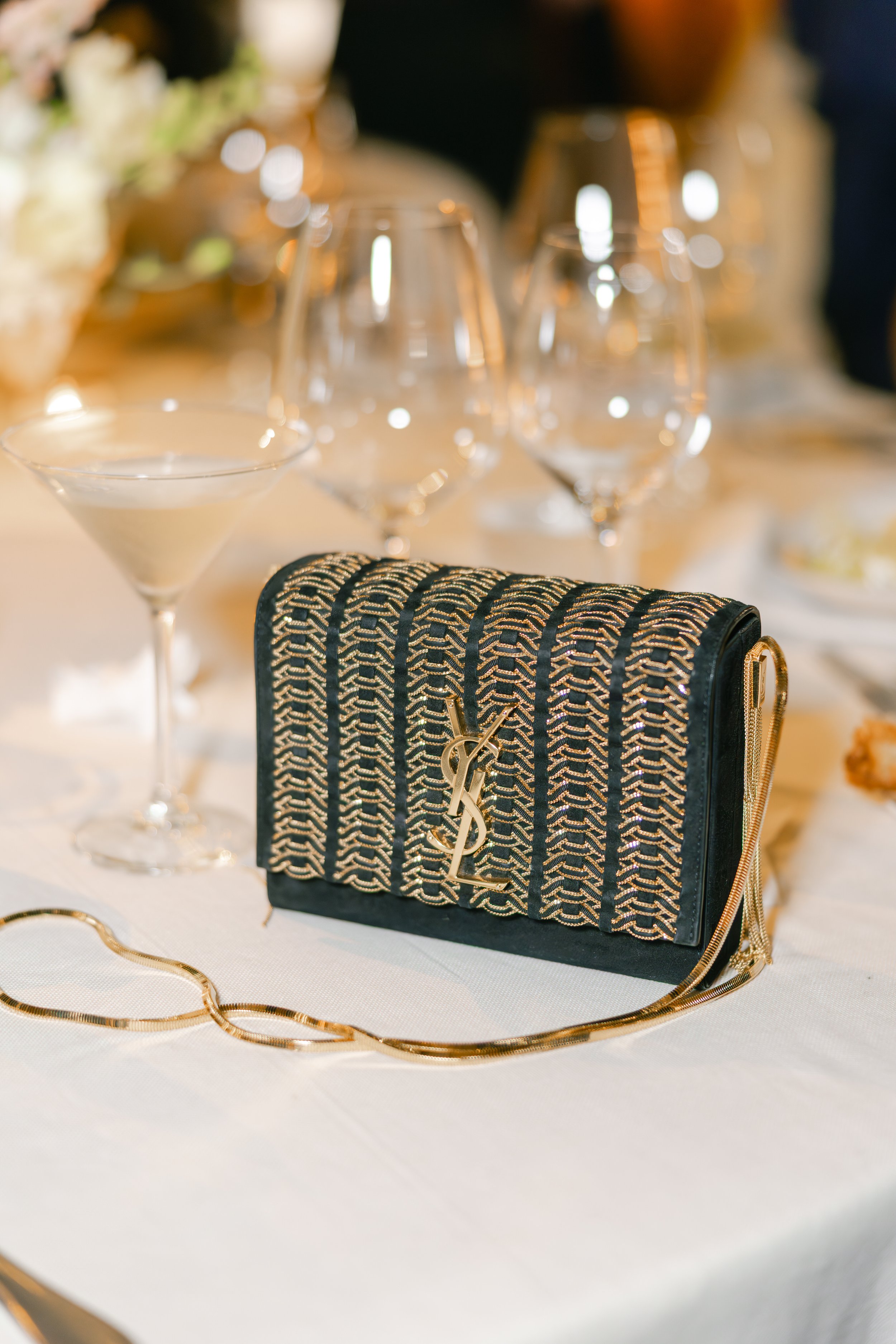 A black designer clutch purse with gold chains and a gold YSL logo on a white tablecloth, surrounded by empty wine glasses and a cocktail glass, at an elegant event or dinner.