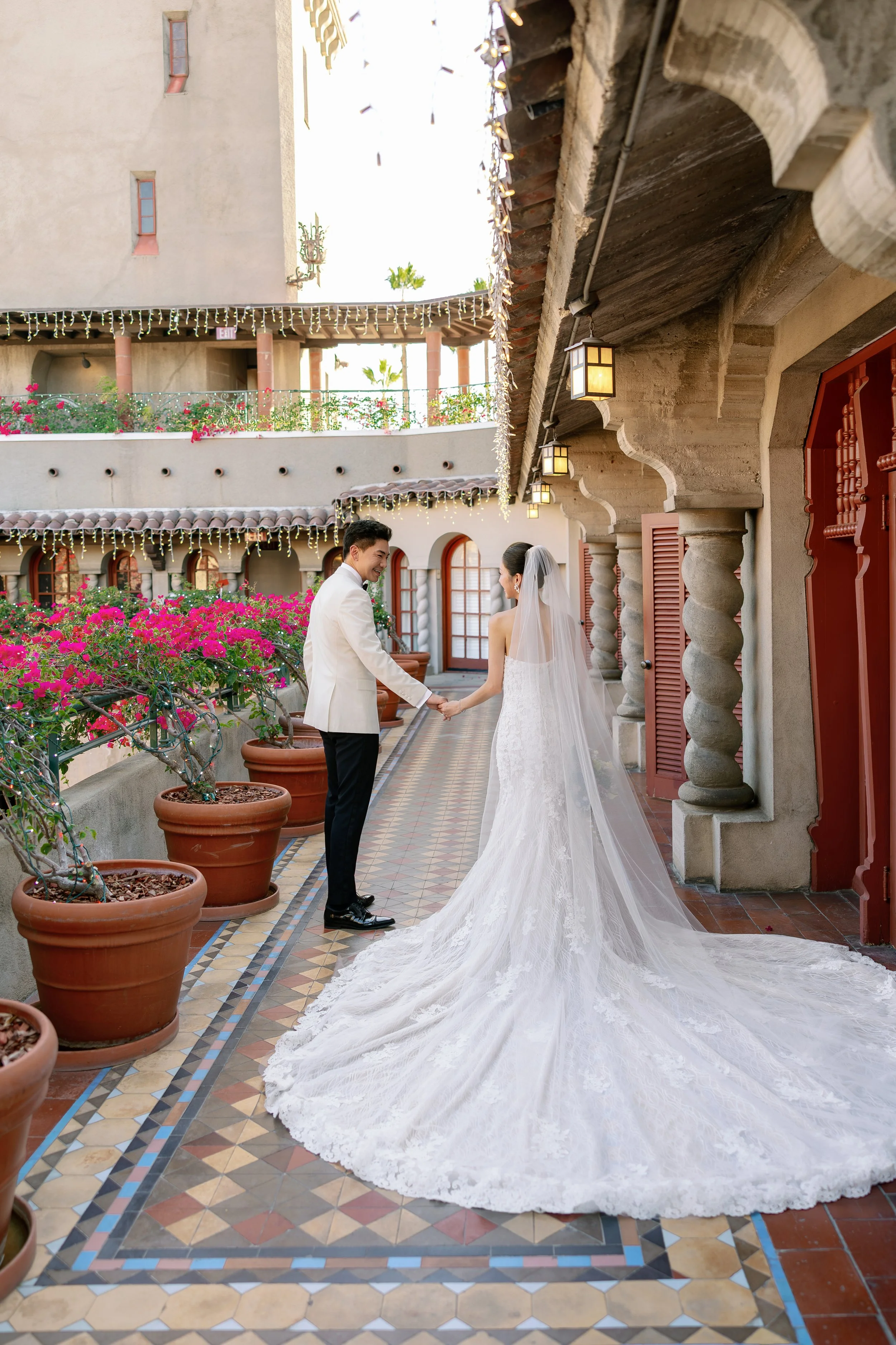 A bride and groom holding hands on a decorative outdoor terrace, with the bride dressed in a white wedding gown with a long train and veil, and the groom in a white suit jacket and black pants, surrounded by potted plants and string lights.