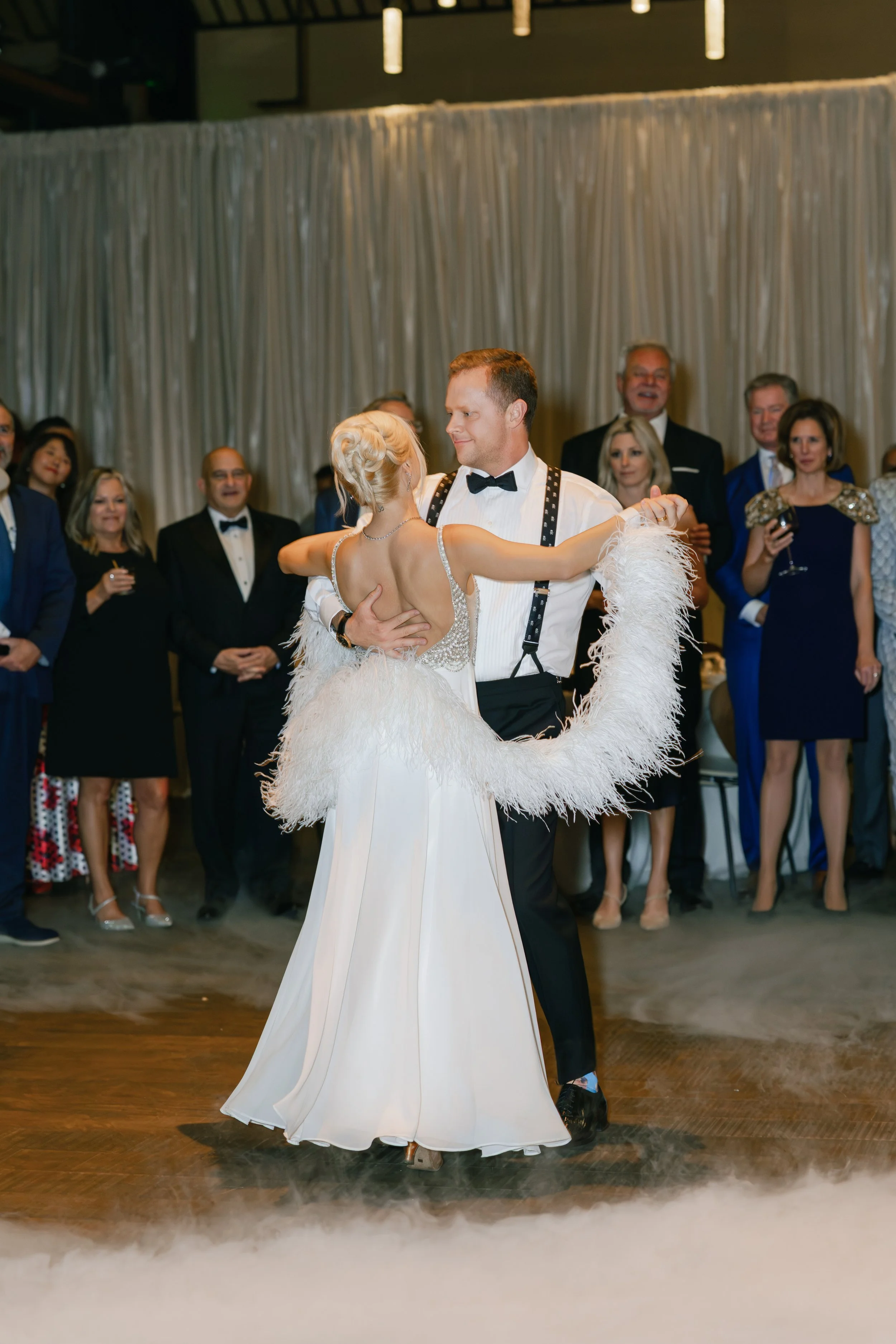 A couple in formal attire dancing at a wedding reception, with guests watching in the background.