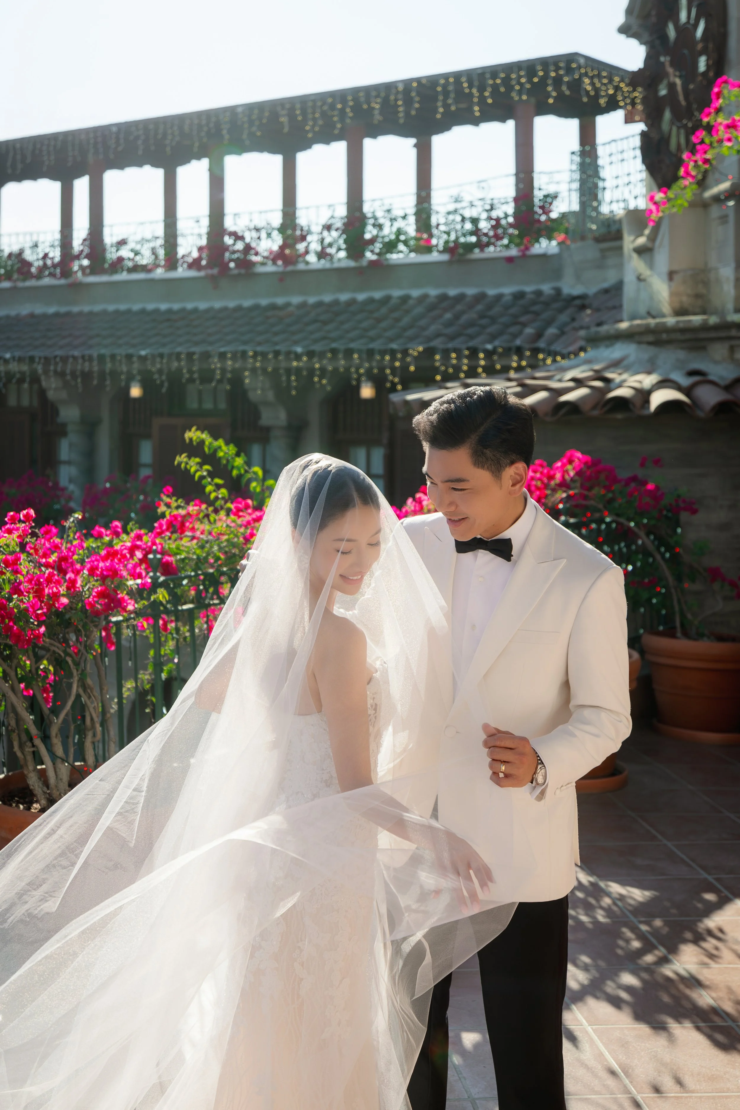 A bride and groom, both smiling, standing on a terrace with pink flowers and string lights, dressed in wedding attire, sharing a moment together.