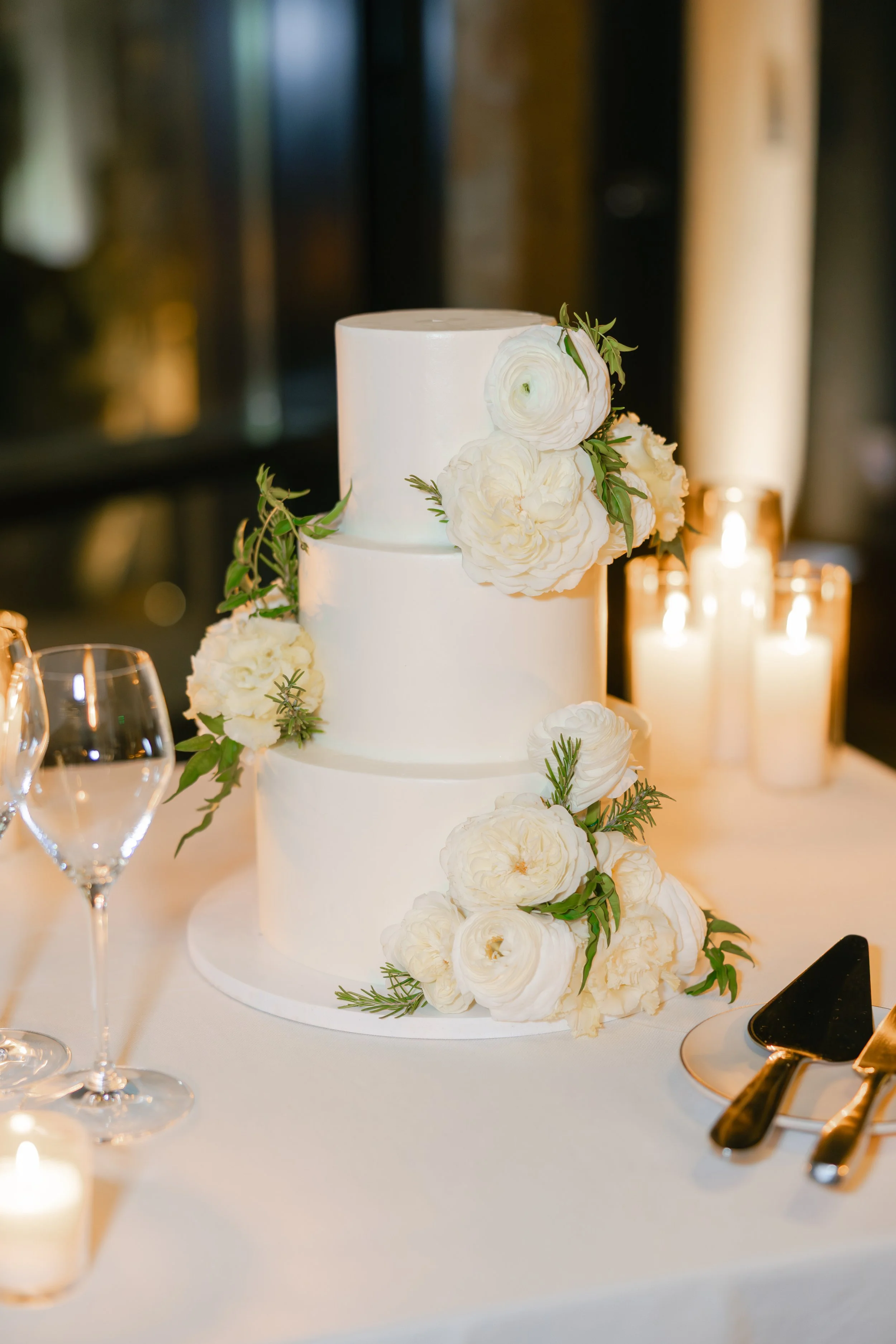 Elegant three-tier white wedding cake decorated with white flowers and greenery, placed on a table with glasses, candles, and silverware.