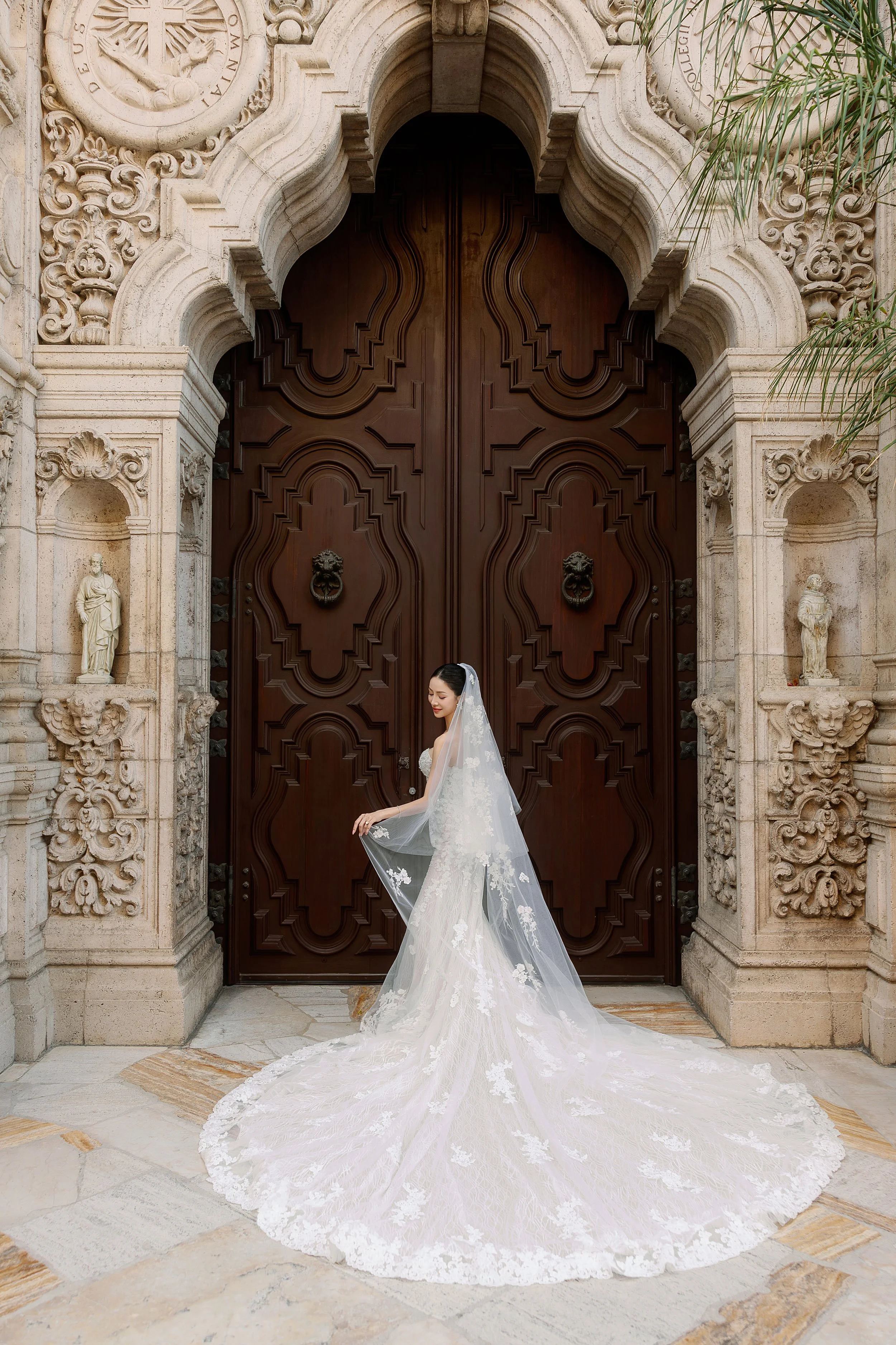 A bride in a white wedding gown with lace details and a long train standing in front of a large, ornate wooden door with detailed carvings and lion head knockers, framed by a decorative stone arch with sculptures and decorative elements.
