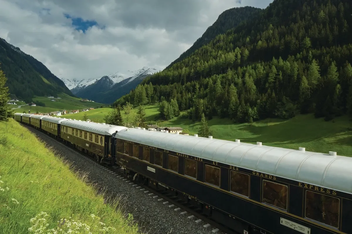 Venice Simplon-Orient-Express luxury train travelling through a dramatic Alpine landscape, with snow-capped mountains and lush green valleys.