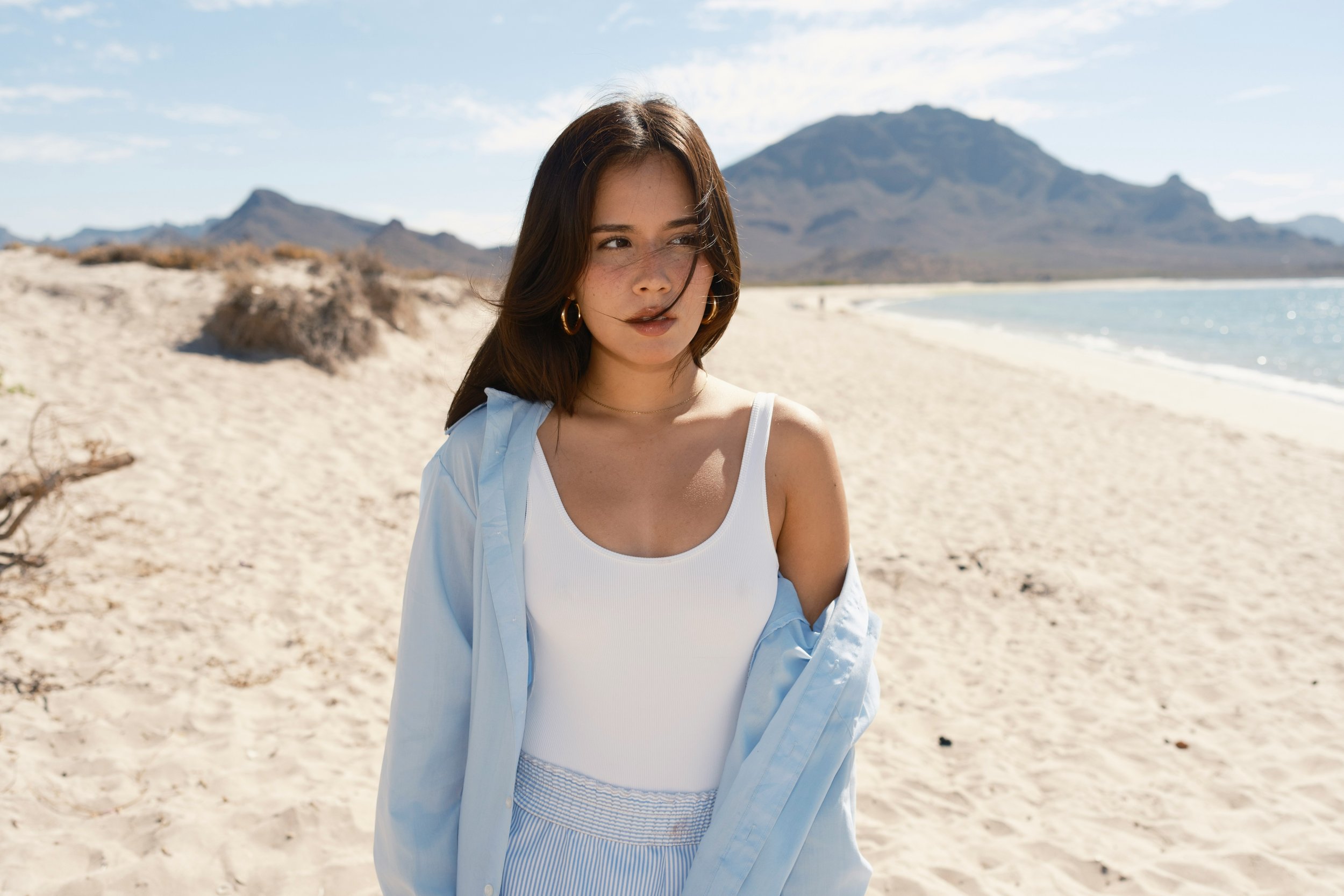 Woman wearing a white swimsuit and pale blue shirt on a sandy beach.