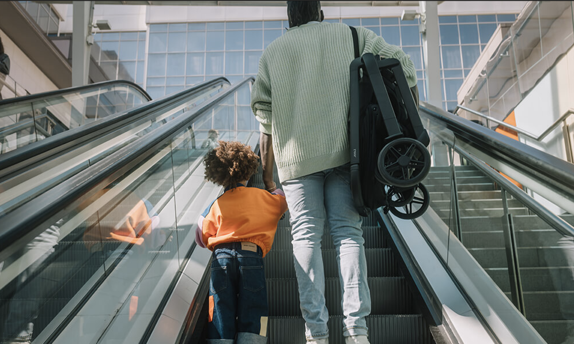 Parent carrying the Joolz Hub2 folded on an escalator with a young child in a busy city setting