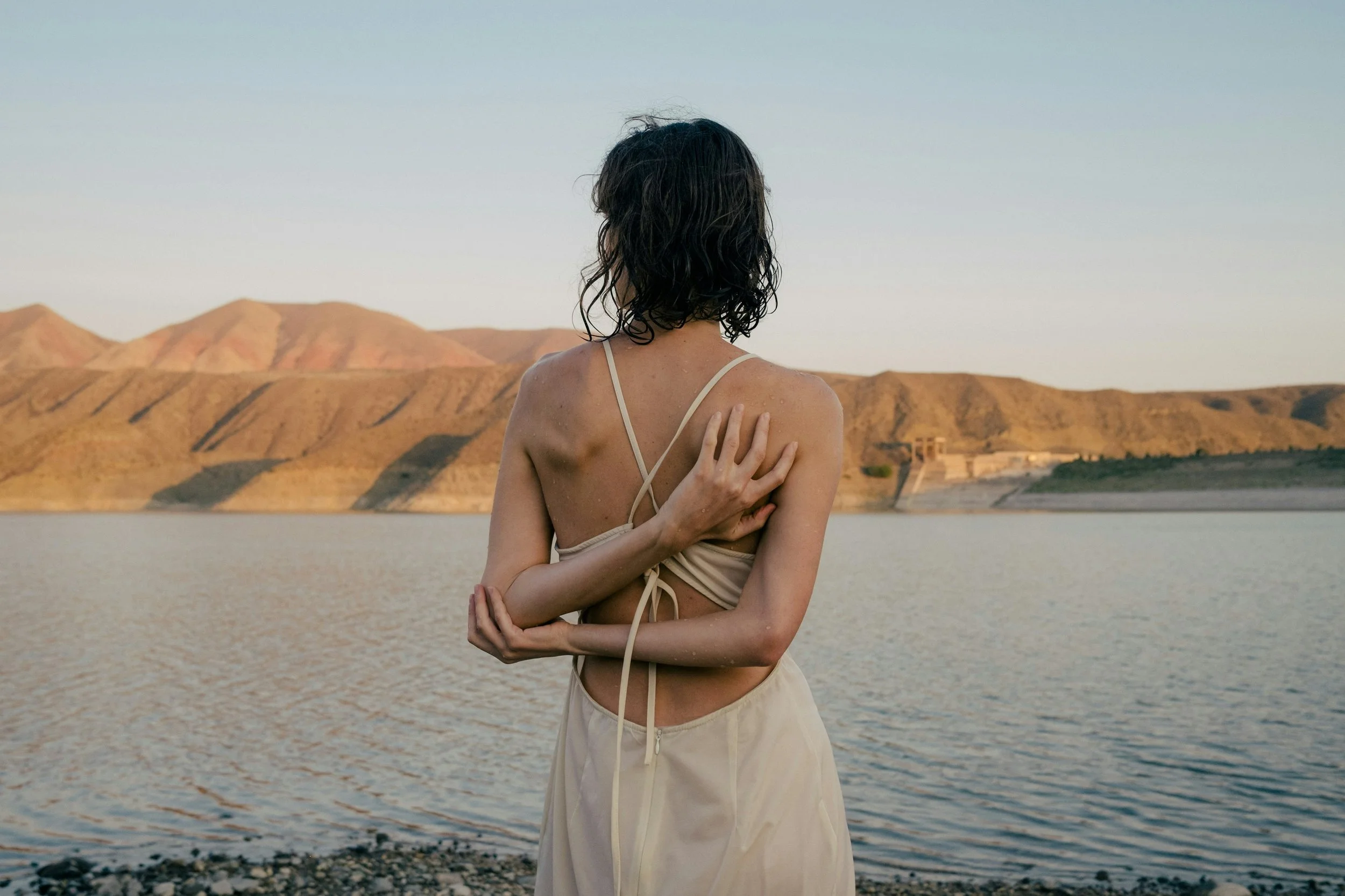 Woman standing by water with her back to the camera, highlighting natural body skin in a calm outdoor setting