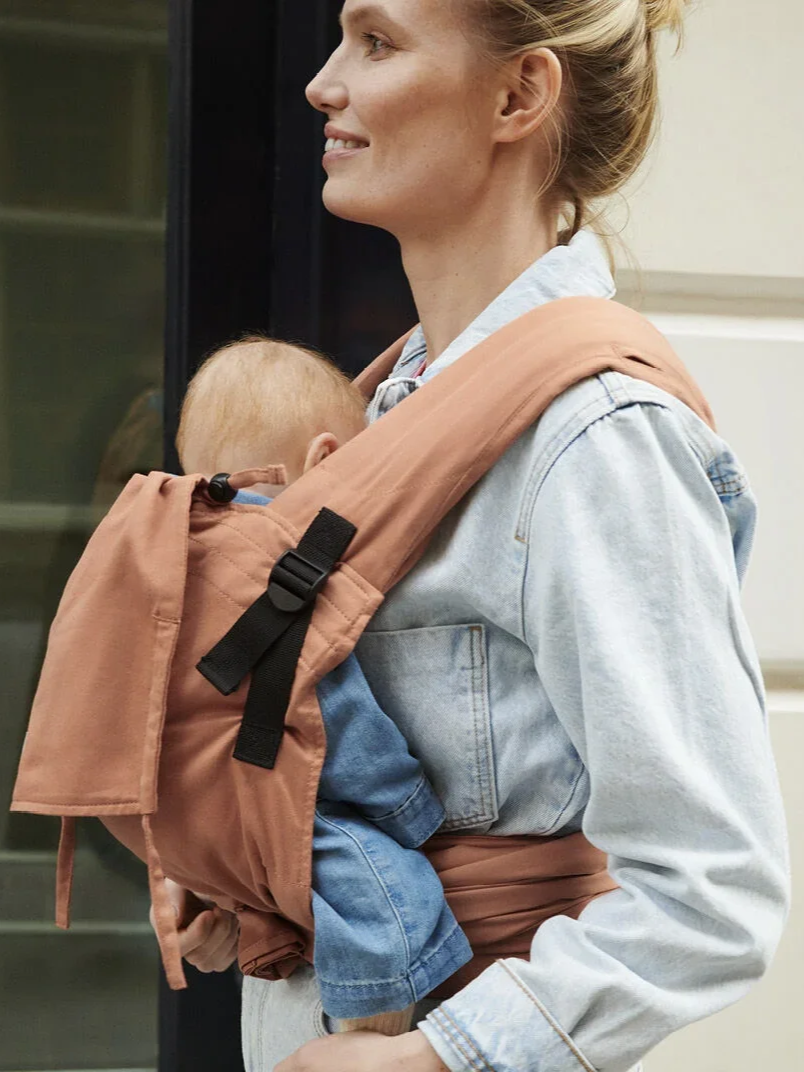 Mother wearing a terracotta Stokke Limas half-buckle baby carrier with her baby