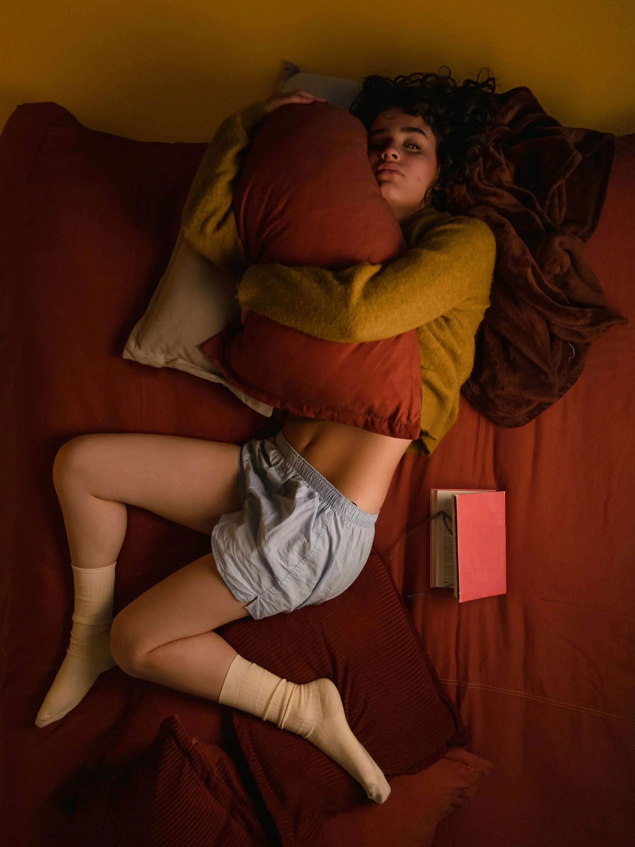 Woman curled up in bed holding a pillow, illustrating fatigue and hormonal sleep disruption linked to perimenopause.