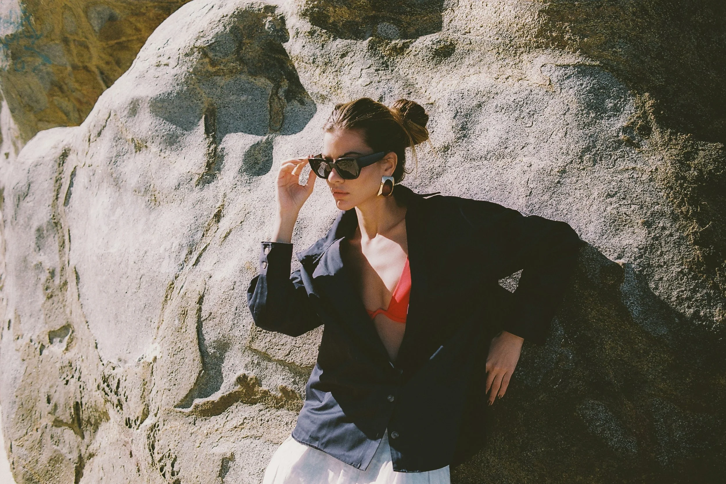 Woman wearing sunglasses leaning against coastal rocks in a luxury summer fashion and beauty editorial