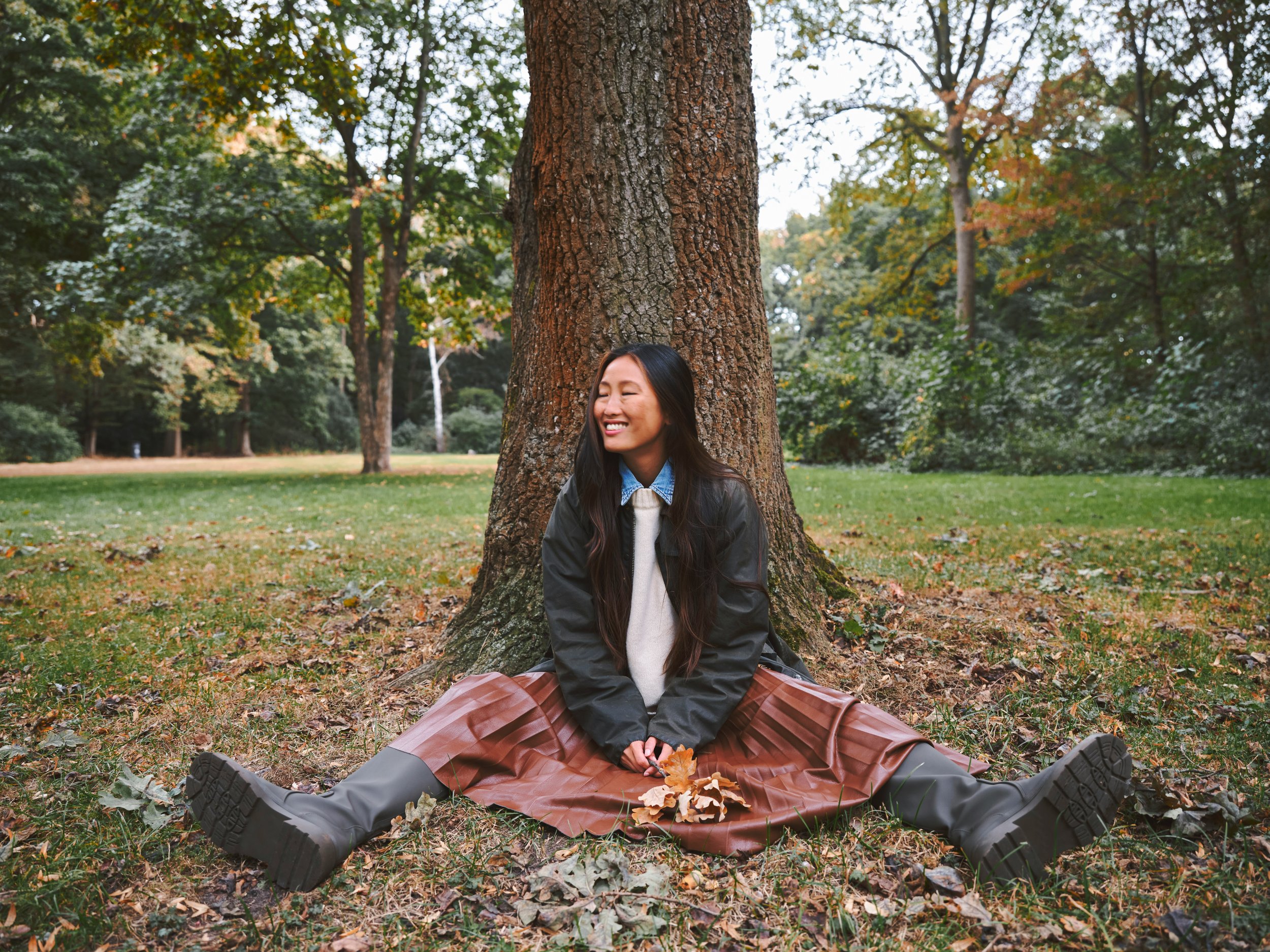 Woman sitting against a tree in a park, smiling naturally and conveying relaxed confidence and effortless style