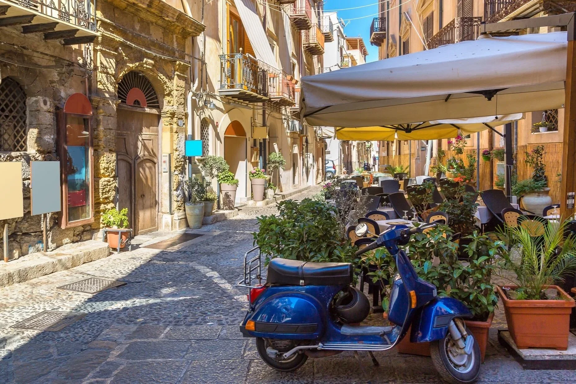 Sicilian street with outdoor café tables and a blue Vespa