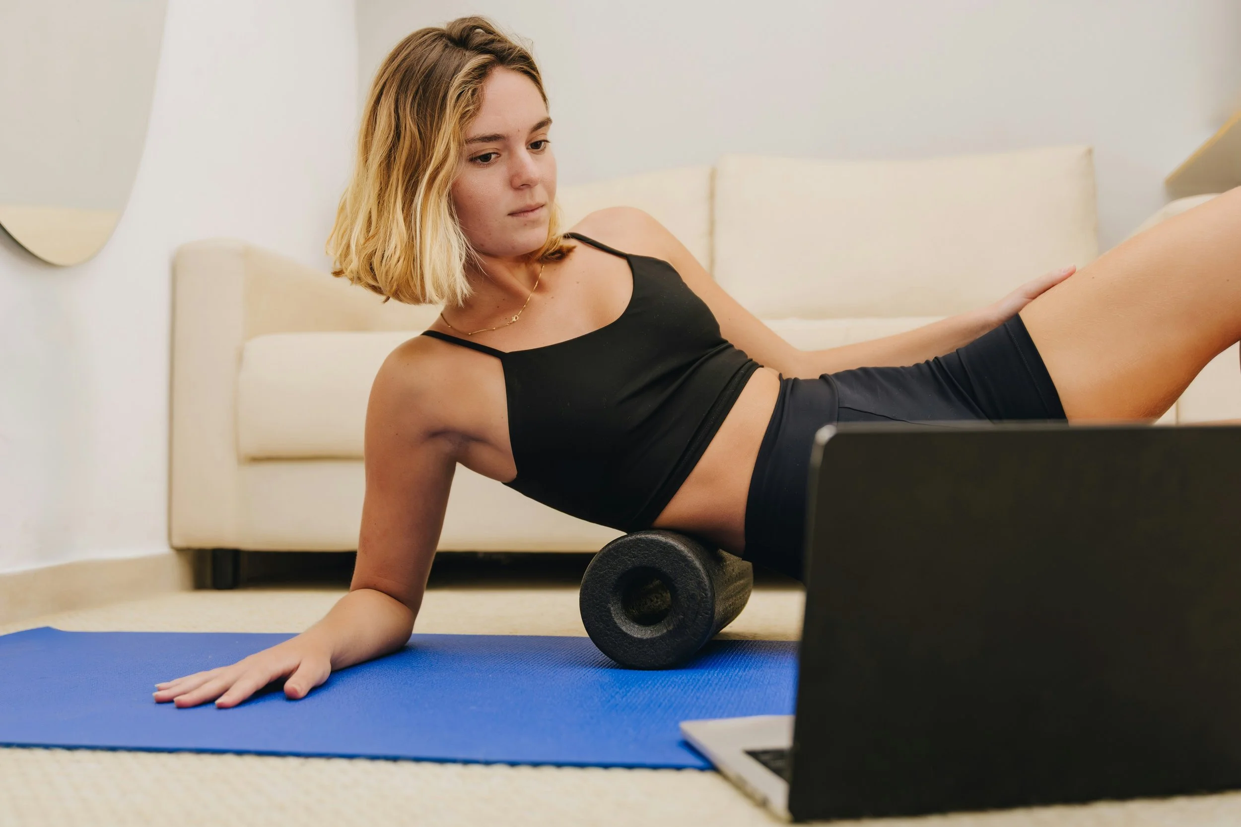 Woman using a foam roller beneath her hip and thigh during a home fitness routine, illustrating self-myofascial release techniques.