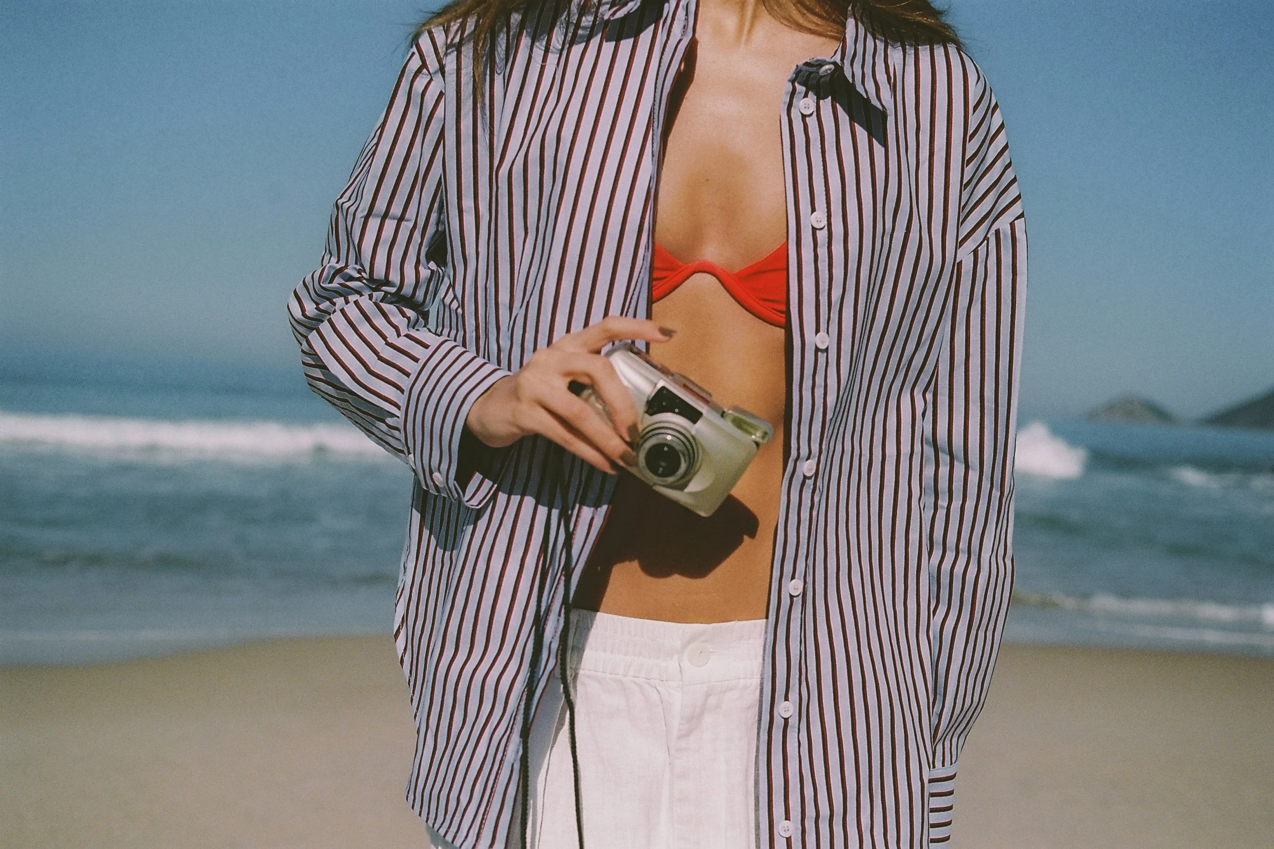 Woman in a striped oversized shirt holding a camera on the beach during a summer editorial shoot