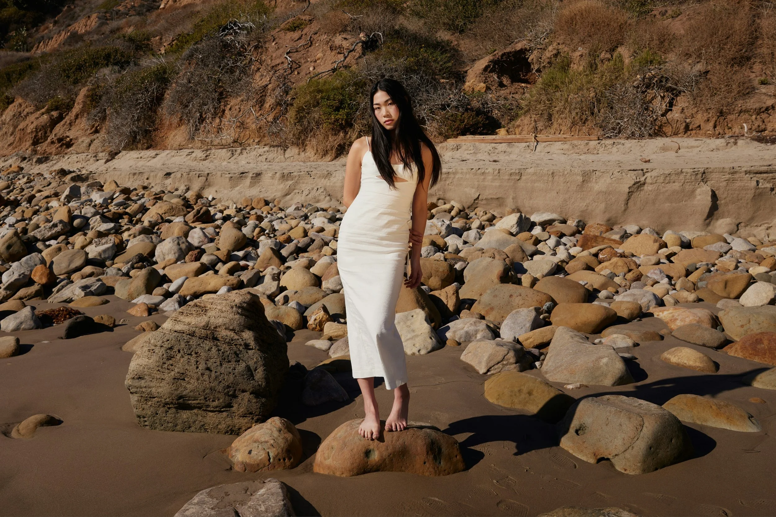 Woman standing barefoot on a rocky beach wearing a white dress, used as editorial imagery for a Morpheus8 skin tightening review