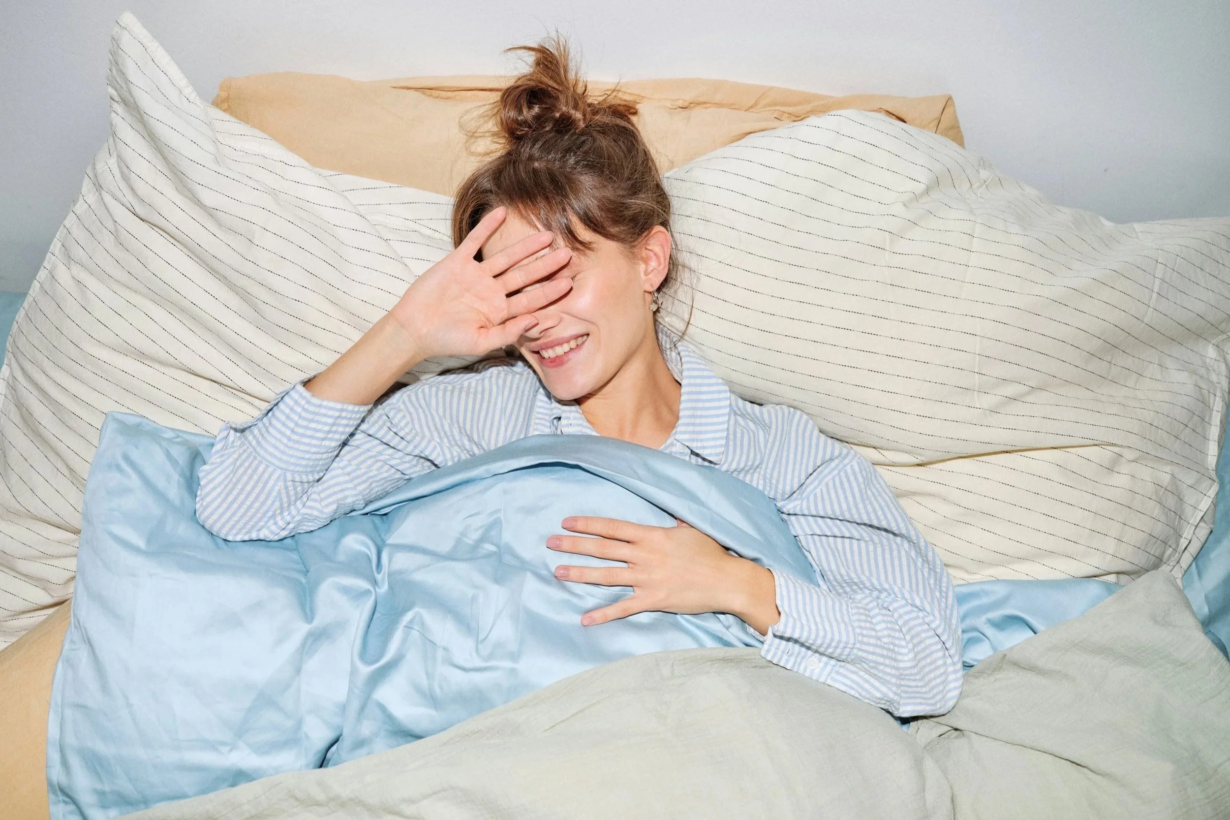Woman lying in bed with one hand covering her face in morning light, capturing a tired, post-celebration moment.
