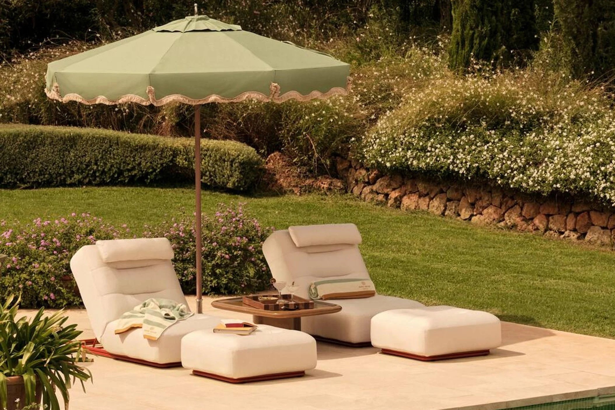 Soho Home Green fringed outdoor parasol beside cream loungers in a Mediterranean-style garden