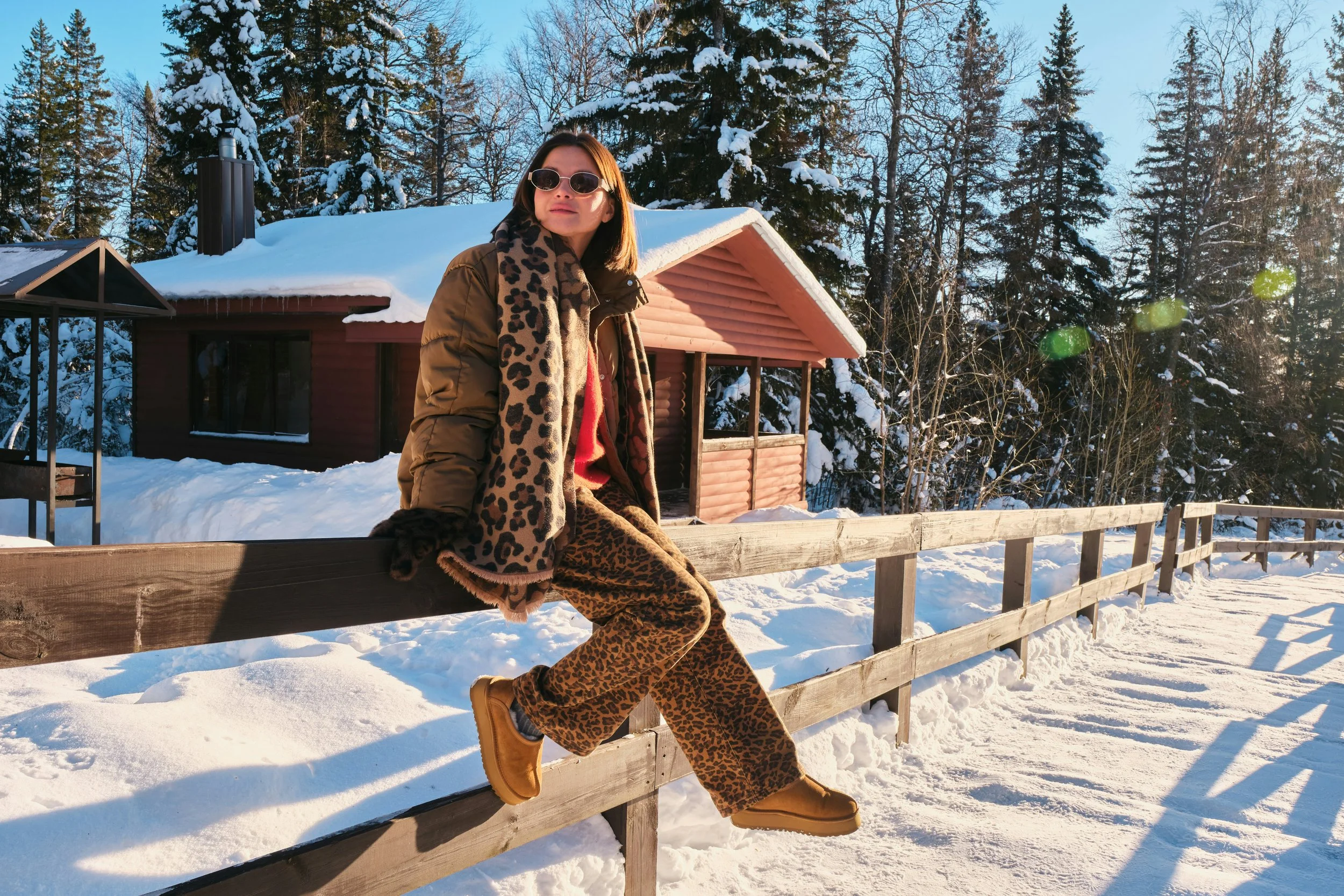 Woman sitting on wooden fence in snowy winter landscape near cabin, dressed in warm clothing