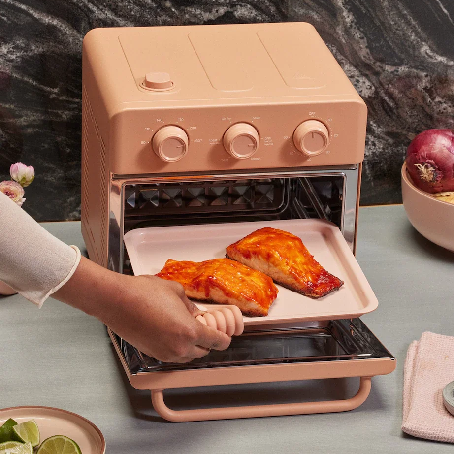 Woman pulls out a tray of salmon steaks after cooking them in a pastel pink Our Place Wonder Oven