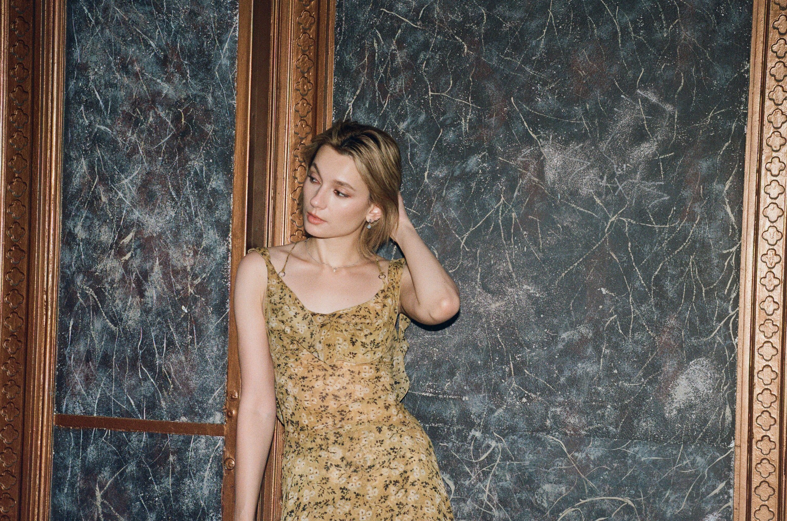 Blonde woman in yellow floral dress leans against wall