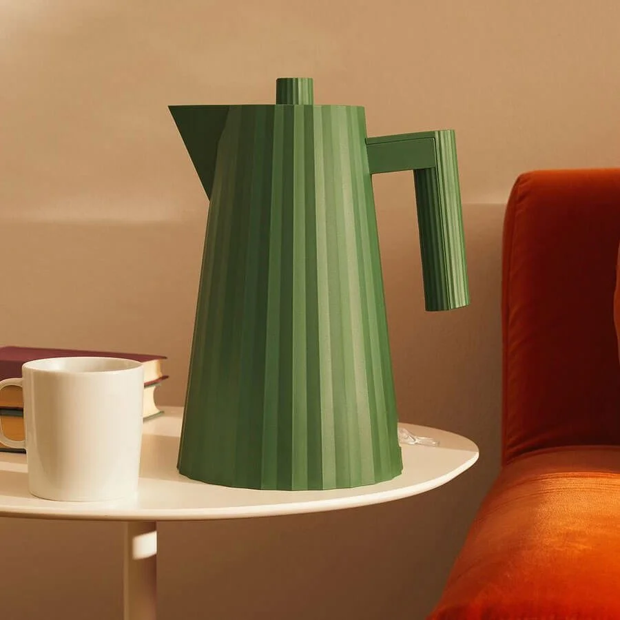 The Alessi Plisse kettle in green sits on a side table next to a cup of coffee.