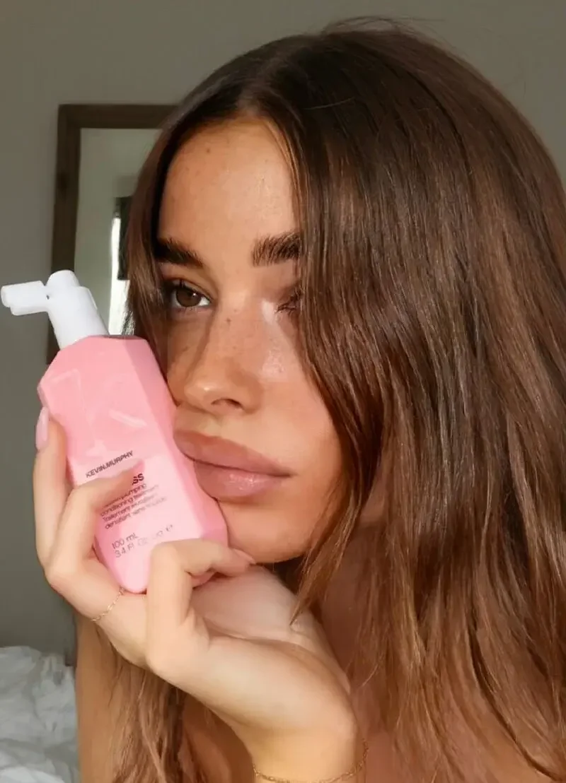 Woman with long, glossy brunette hair holding a pink Kevin Murphy haircare bottle beside her face.