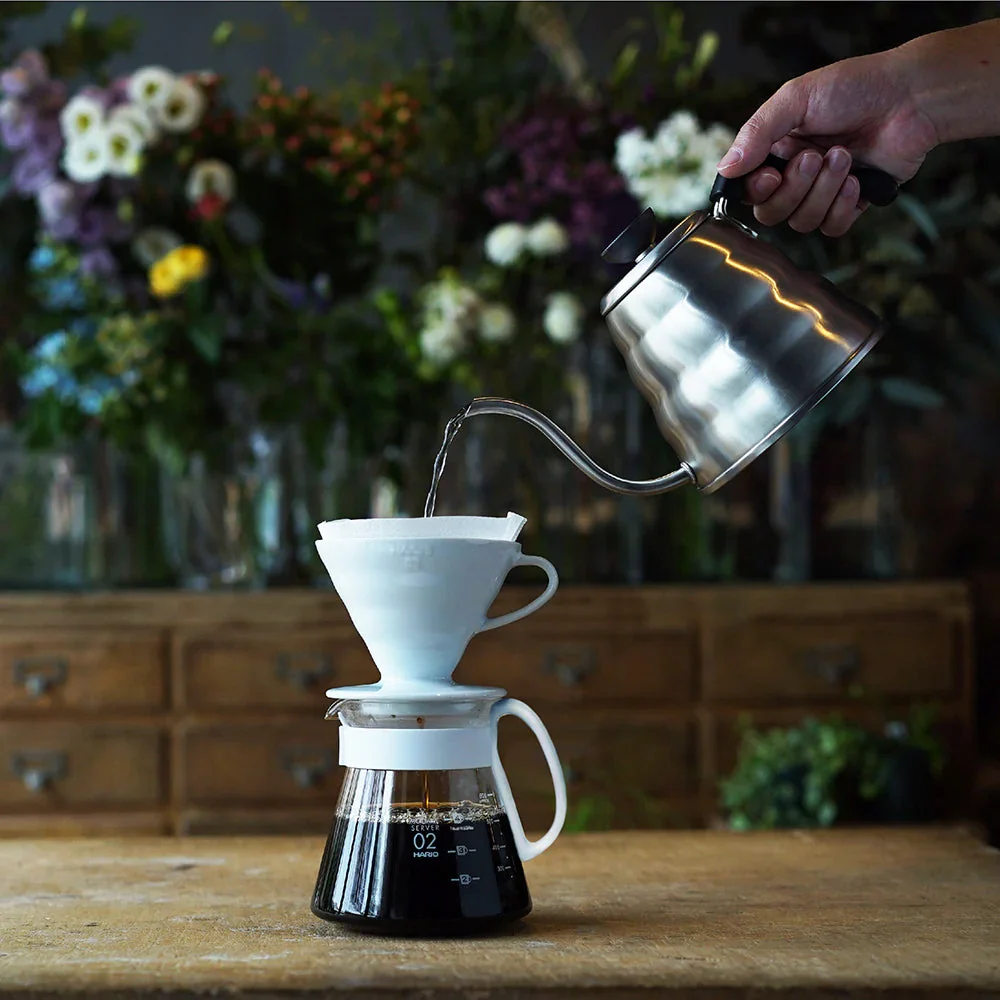 Boiling water poured from the Hario Buono Kettle into the Hario Ceramic Coffee dripper in front of a table full of flowers