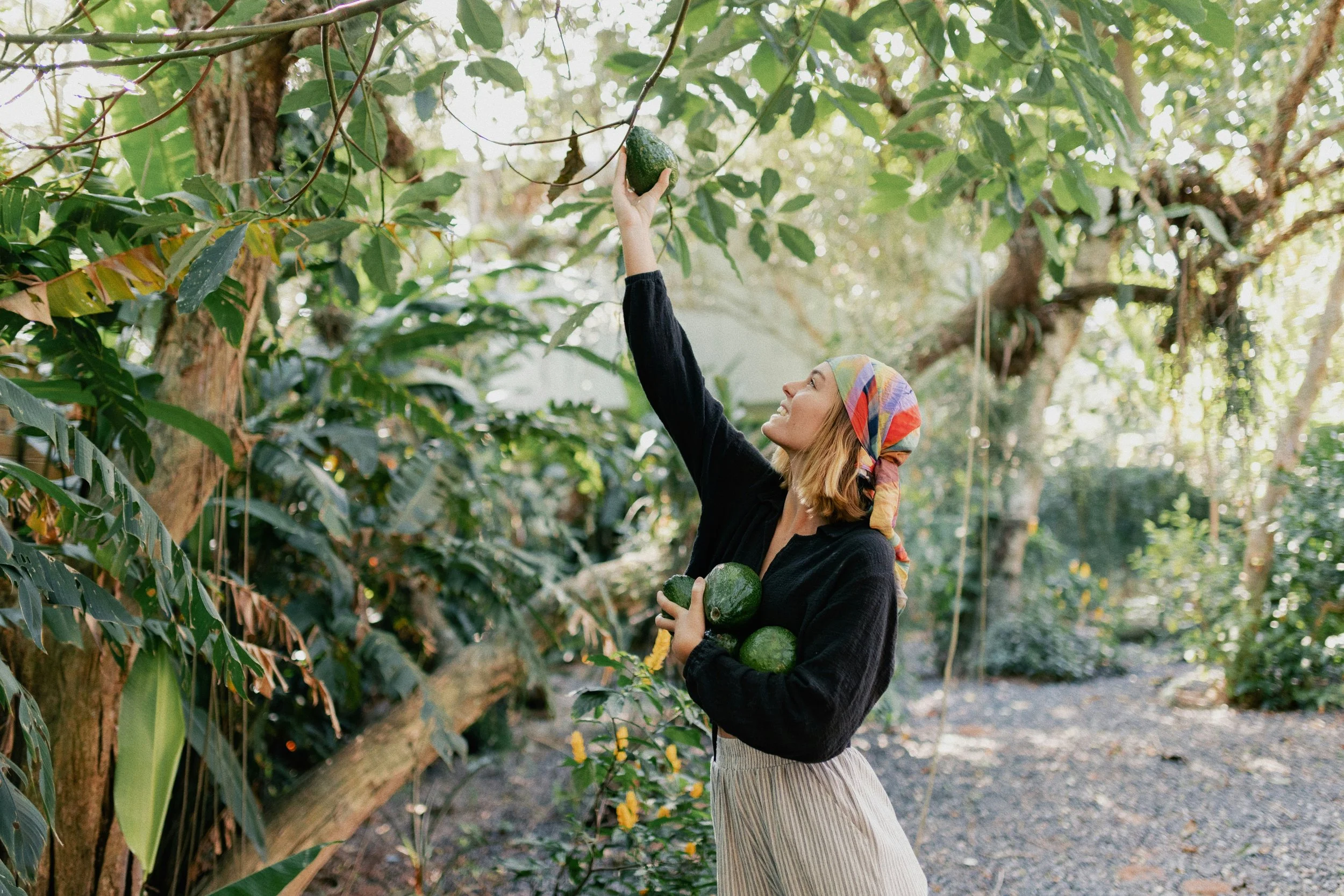 t: Woman reaching into an avocado tree in relaxed, timeless clothing, representing the natural evolution of personal style.