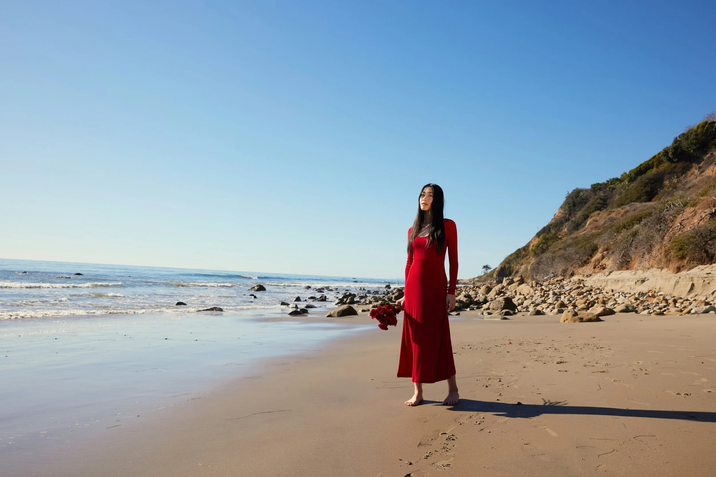 woman weaing red dress, hold a bunch of flowers in her hands while walking bare feet on the beach