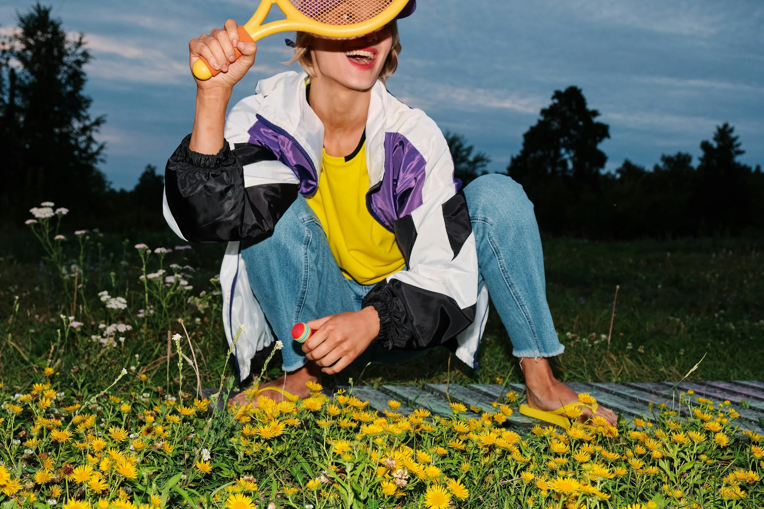 Woman crouching outdoors at dusk holding a yellow racket in a wildflower meadow