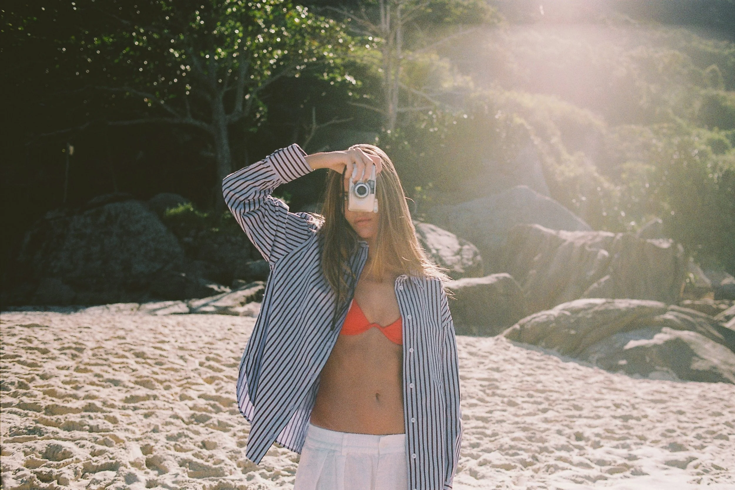 Woman holding a camera on a beach wearing a striped shirt and red bikini for a summer beauty editorial