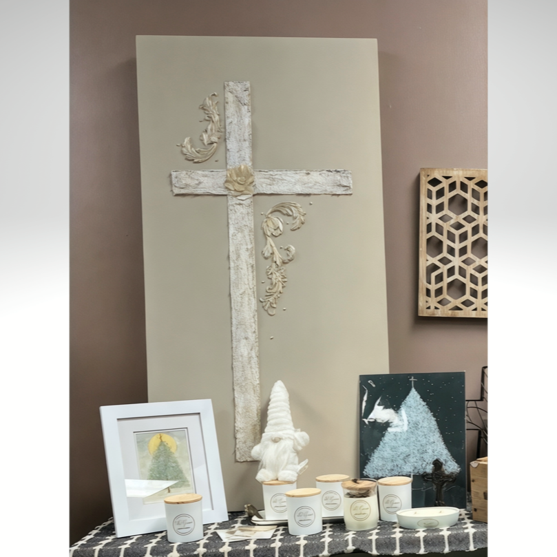 Decorative display featuring a white cross with ornate embellishments on a beige background, surrounded by framed artwork, candles, and decorative objects.