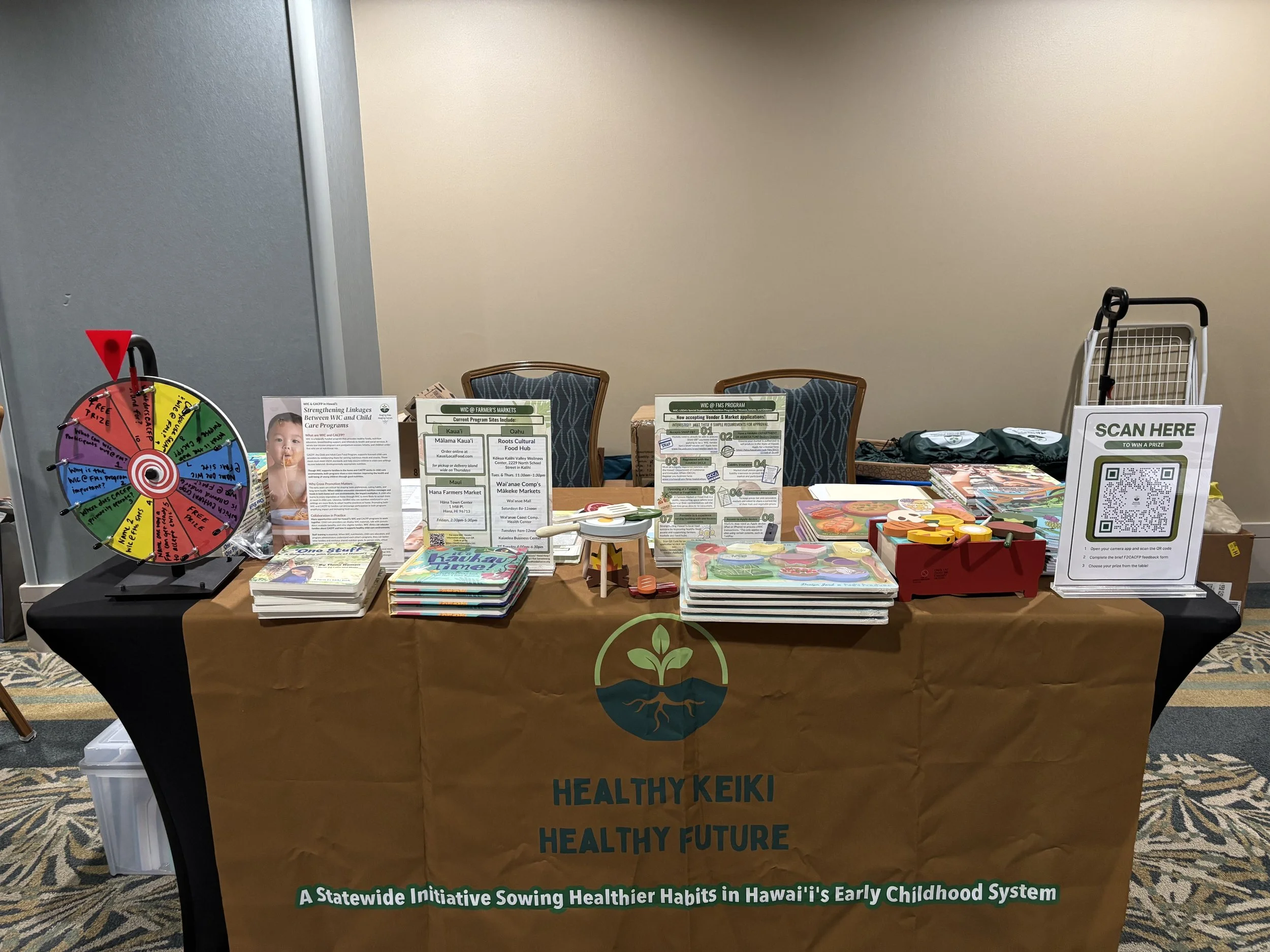 A booth at an event displaying informational material, a prize wheel, books, and a sign that reads 'SCAN HERE TO WIN A PRIZE.' The tablecloth has the logo of a tree with roots and the words 'HEALTHY KEIKI, HEALTHY FUTURE.' The display promotes a stat