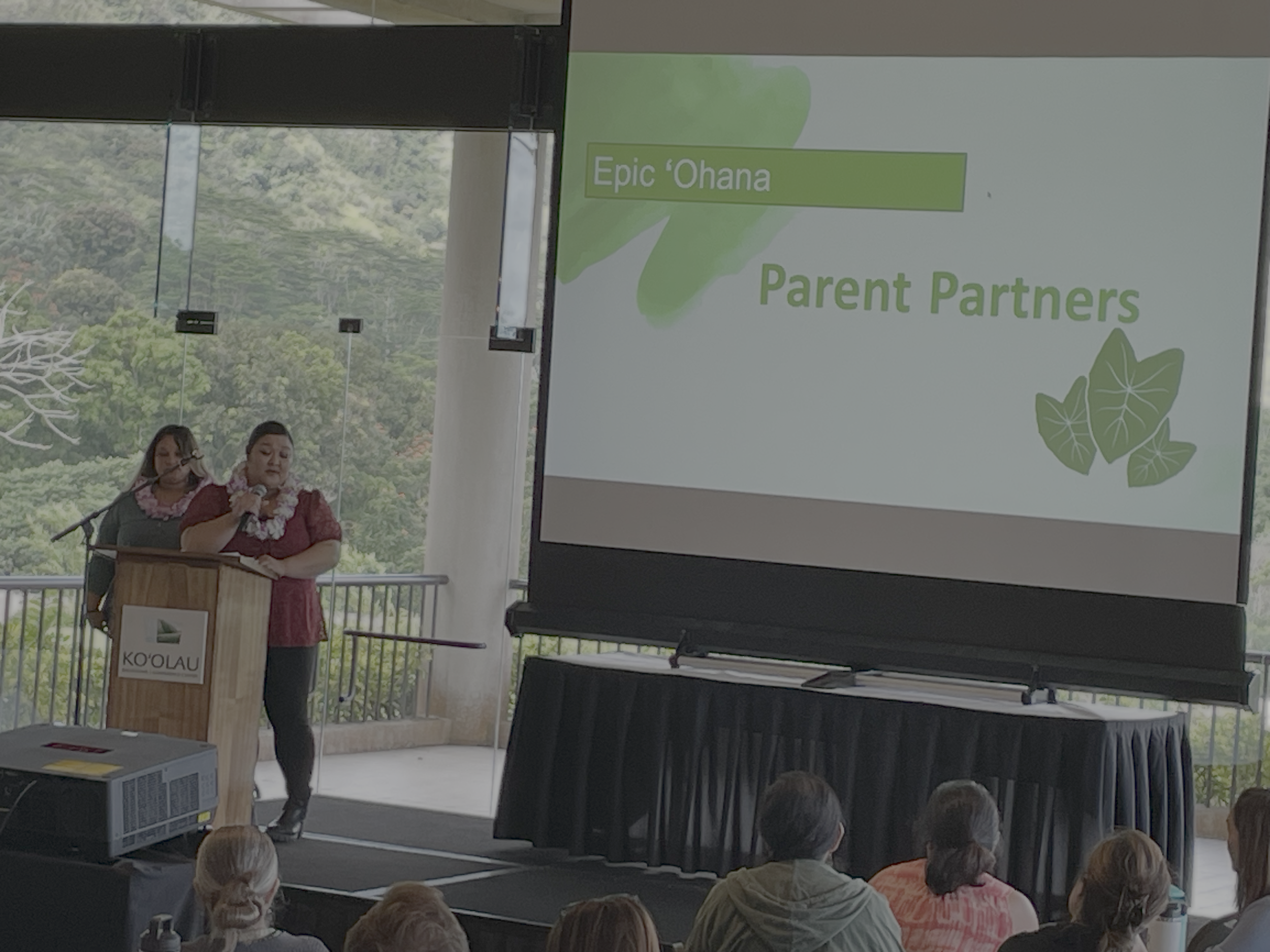 A speaker at a podium giving a presentation at an indoor event, with a large screen displaying the title 'Epic 'Ohana' and 'Parent Partners', and an audience seated in front.