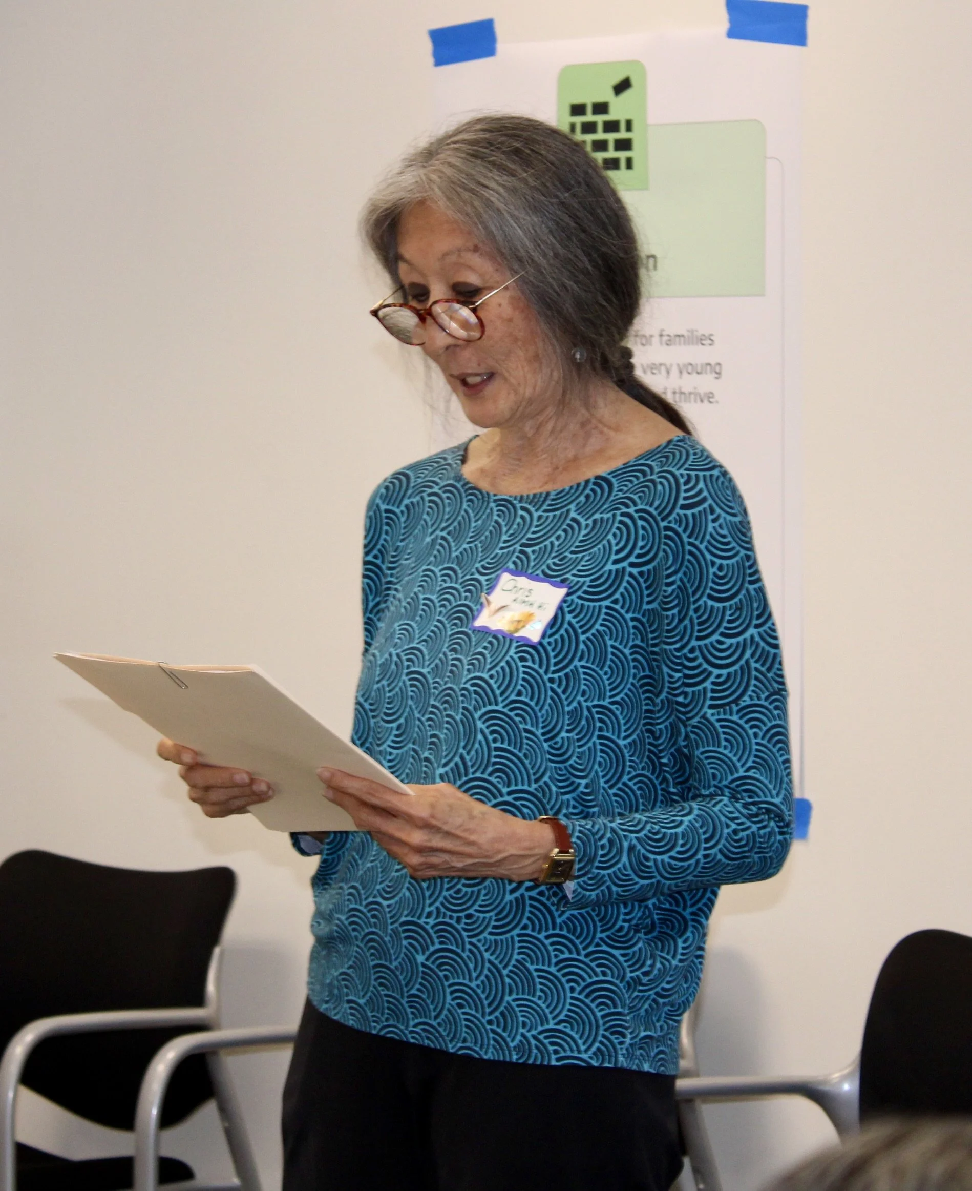 A woman with gray hair wearing glasses on the tip of her nose, dressed in a blue patterned top, standing and reading from a paper or book in a conference room with black chairs in the background.