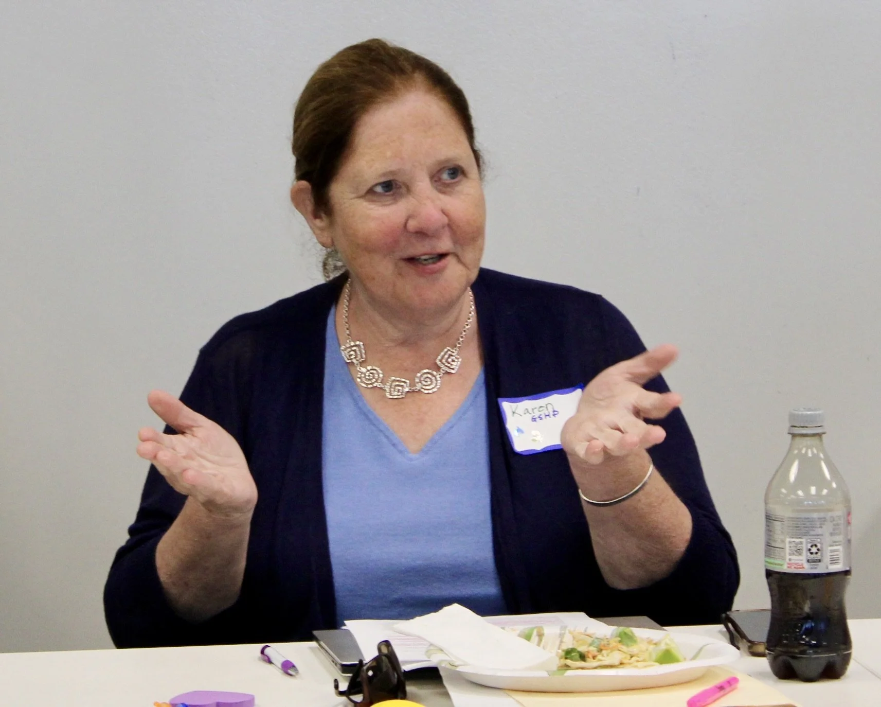 A woman wearing a blue shirt and black cardigan, with a name tag that reads 'Karen,' is sitting at a table with a plate of food, a bottle of soda, and various markers and papers, engaging in conversation.