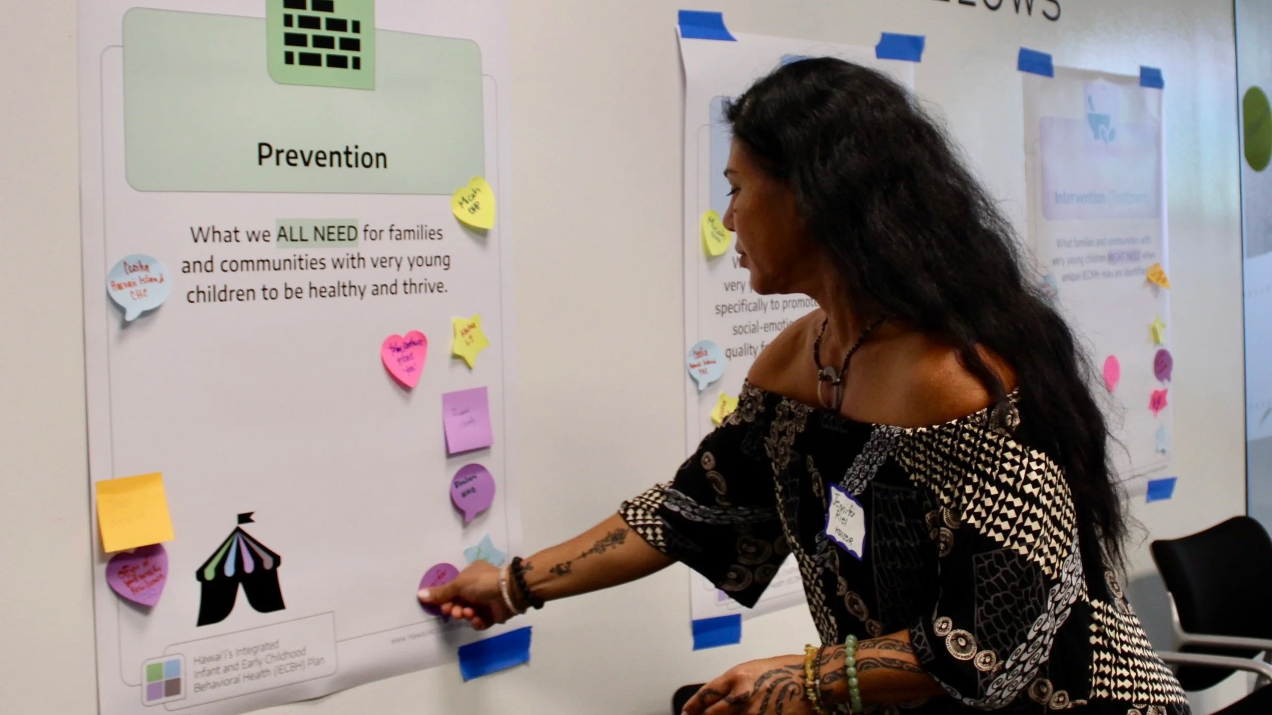 A woman with long dark curly hair in a patterned black and white shirt is taping colorful sticky notes on a whiteboard that contains informational posters and diagrams about prevention and early childhood health.
