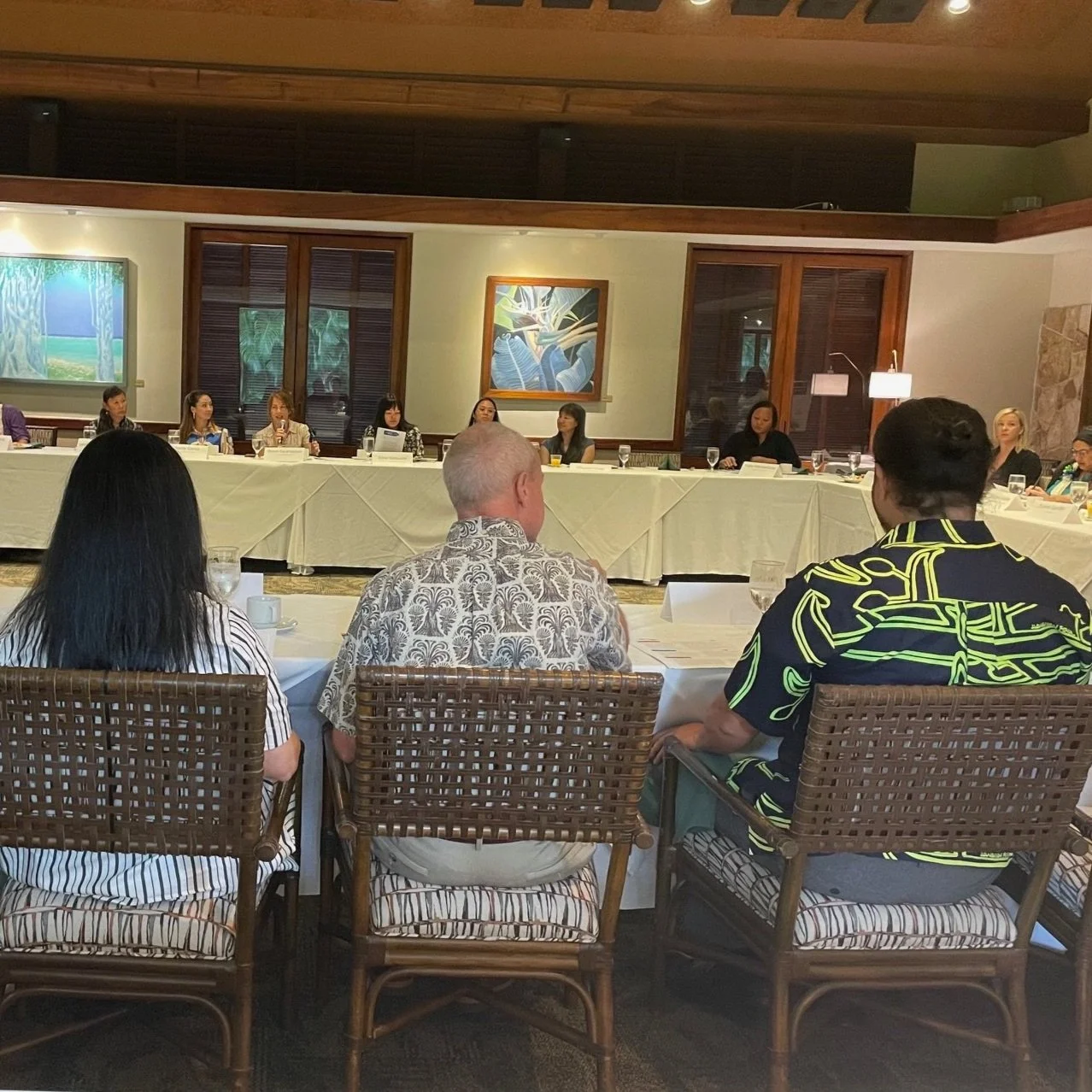 People sitting around a banquet table in a conference room, with a long head table at the front and artwork on the walls.