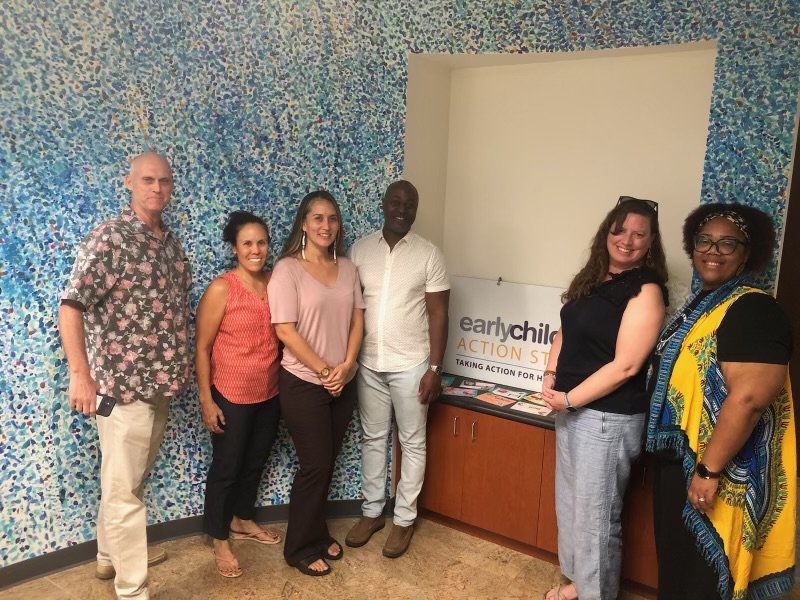 Six people standing together indoors, posing for a photo in front of a blue mosaic wall and a sign that reads 'earlychild ACTION' with additional text underneath.