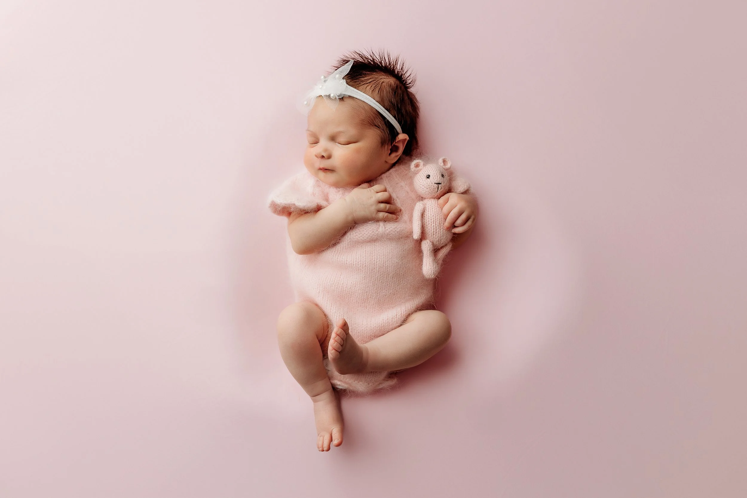 A sleeping baby with dark hair, wearing a pink outfit and headband with a bow, lying on a pink background holding a pink knitted teddy bear.