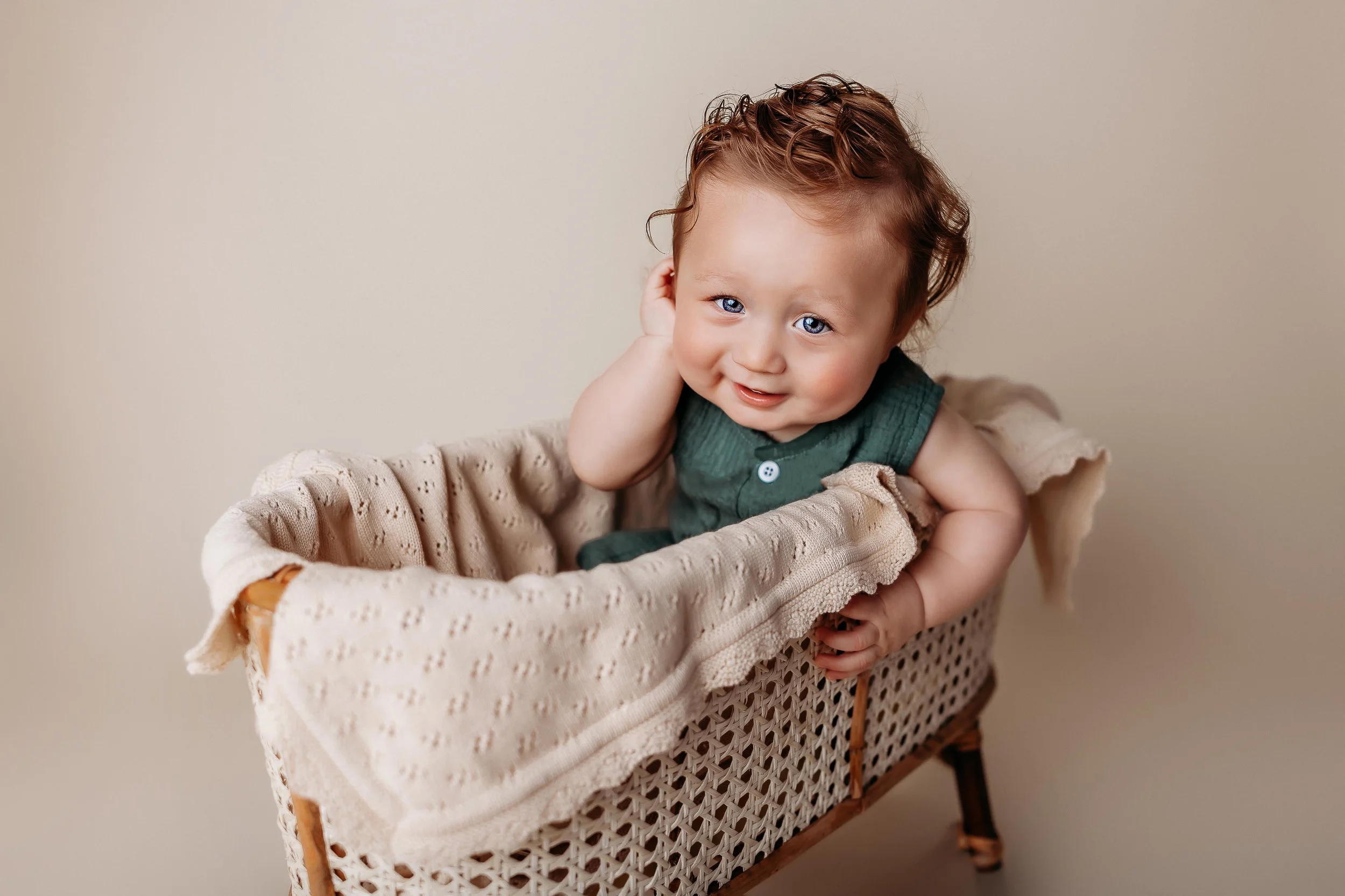A smiling baby with curly brown hair and blue eyes sitting in a woven bassinet lined with a beige blanket, wearing a dark green sleeveless top, holding one hand to their ear, against a plain light background.