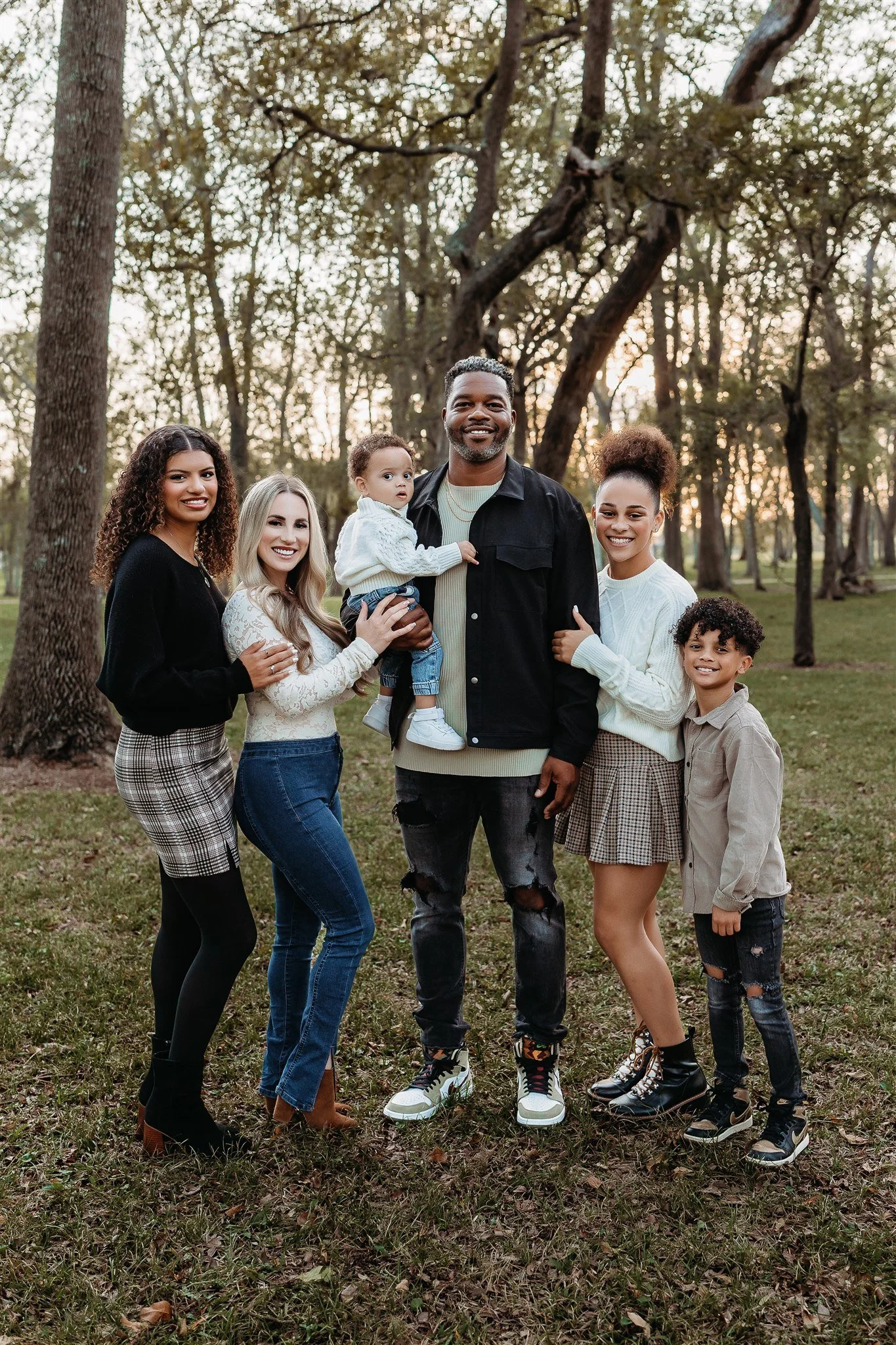 A diverse group of seven people, including children and adults, standing outdoors in a wooded park during sunset, smiling at the camera.