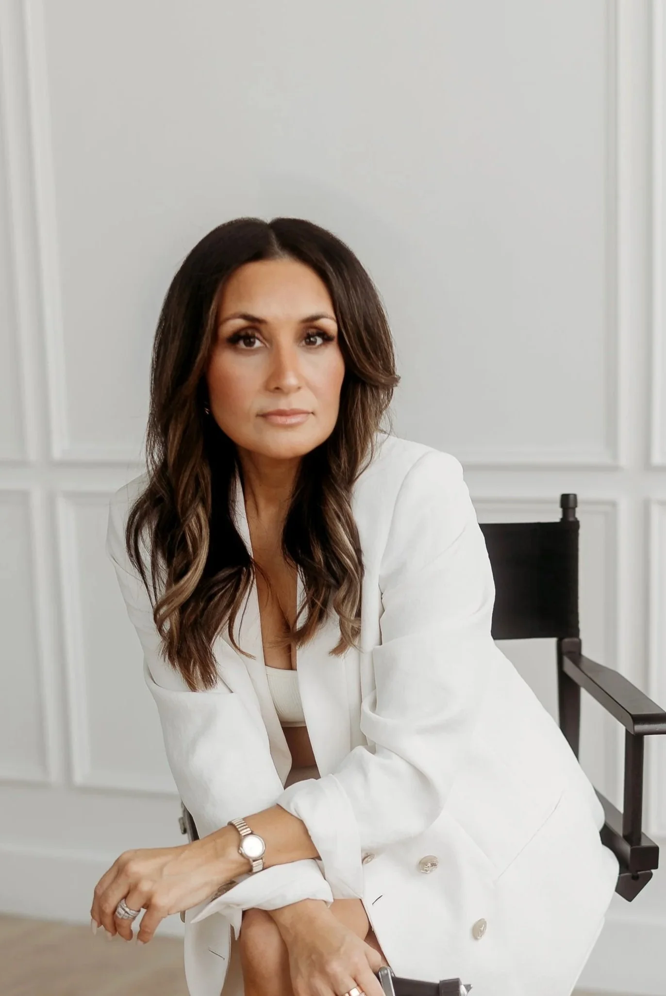 A woman with long wavy brown hair sitting in a black director's chair in front of a white wall, wearing a white blazer, white dress, and a silver watch.