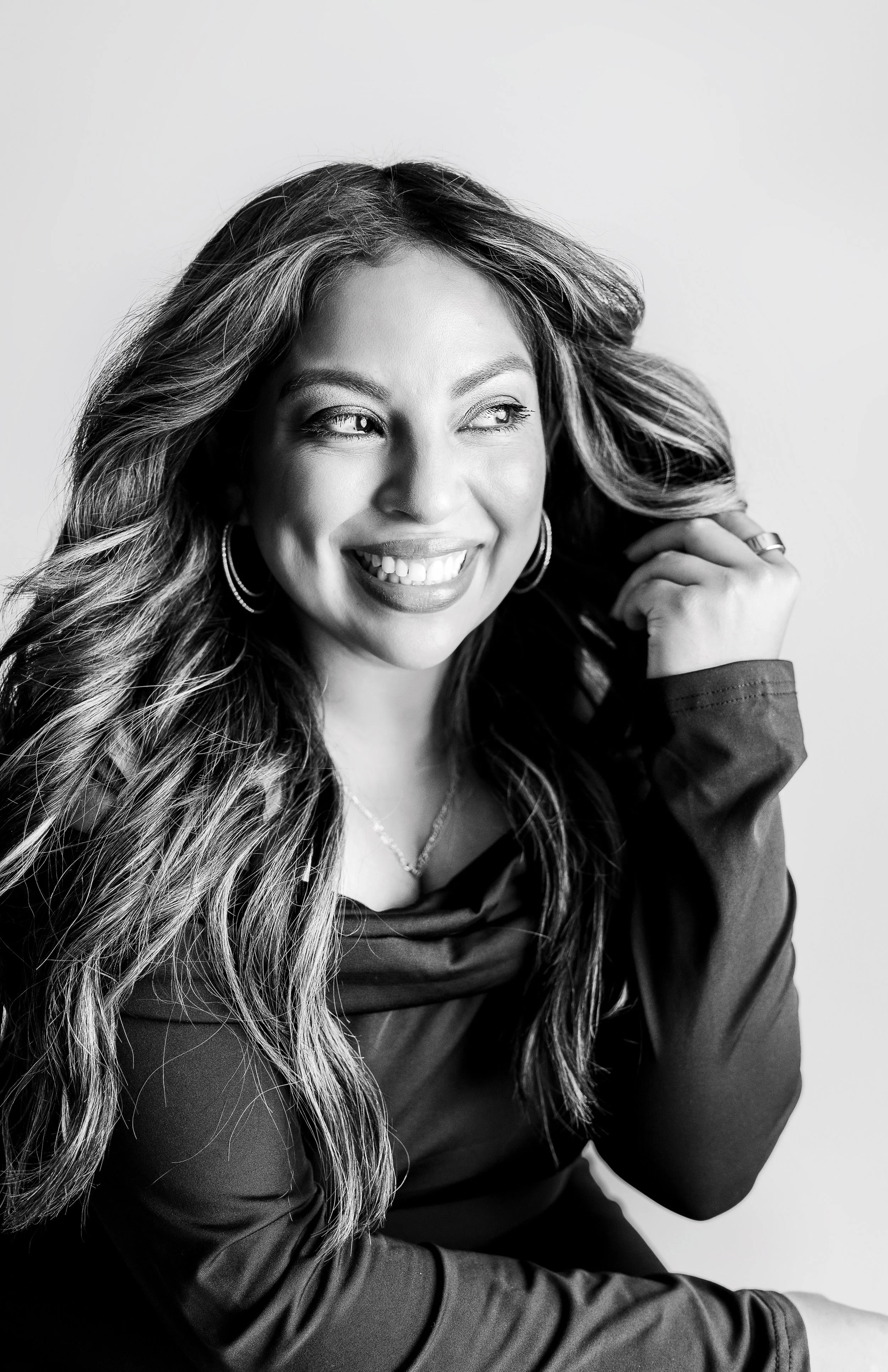Black and white portrait of a woman smiling, with long wavy hair, hoop earrings, and a black jacket, sitting against a plain background.