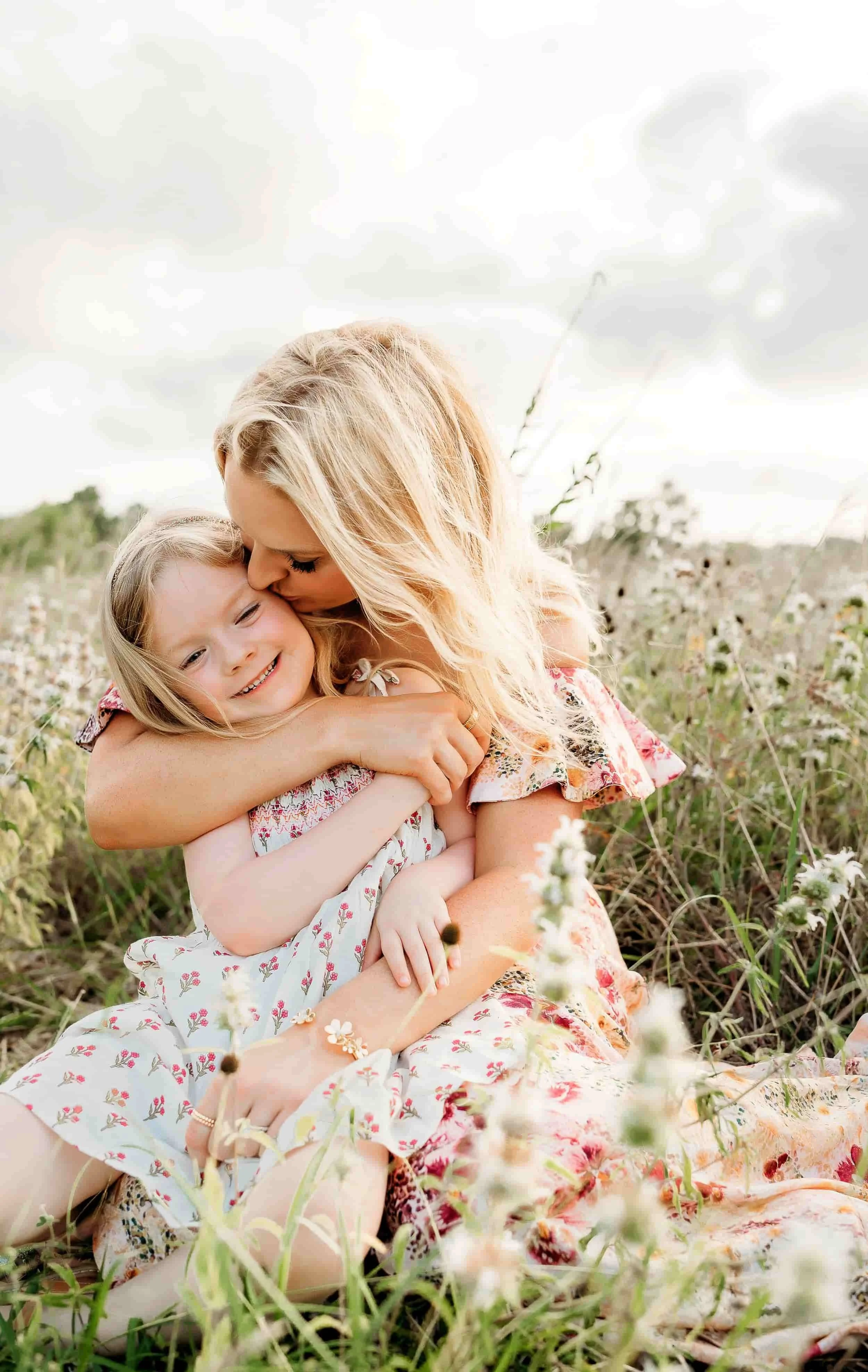 A woman with long blonde hair and a young girl with blonde hair hugging each other in a field of white flowers under a cloudy sky.